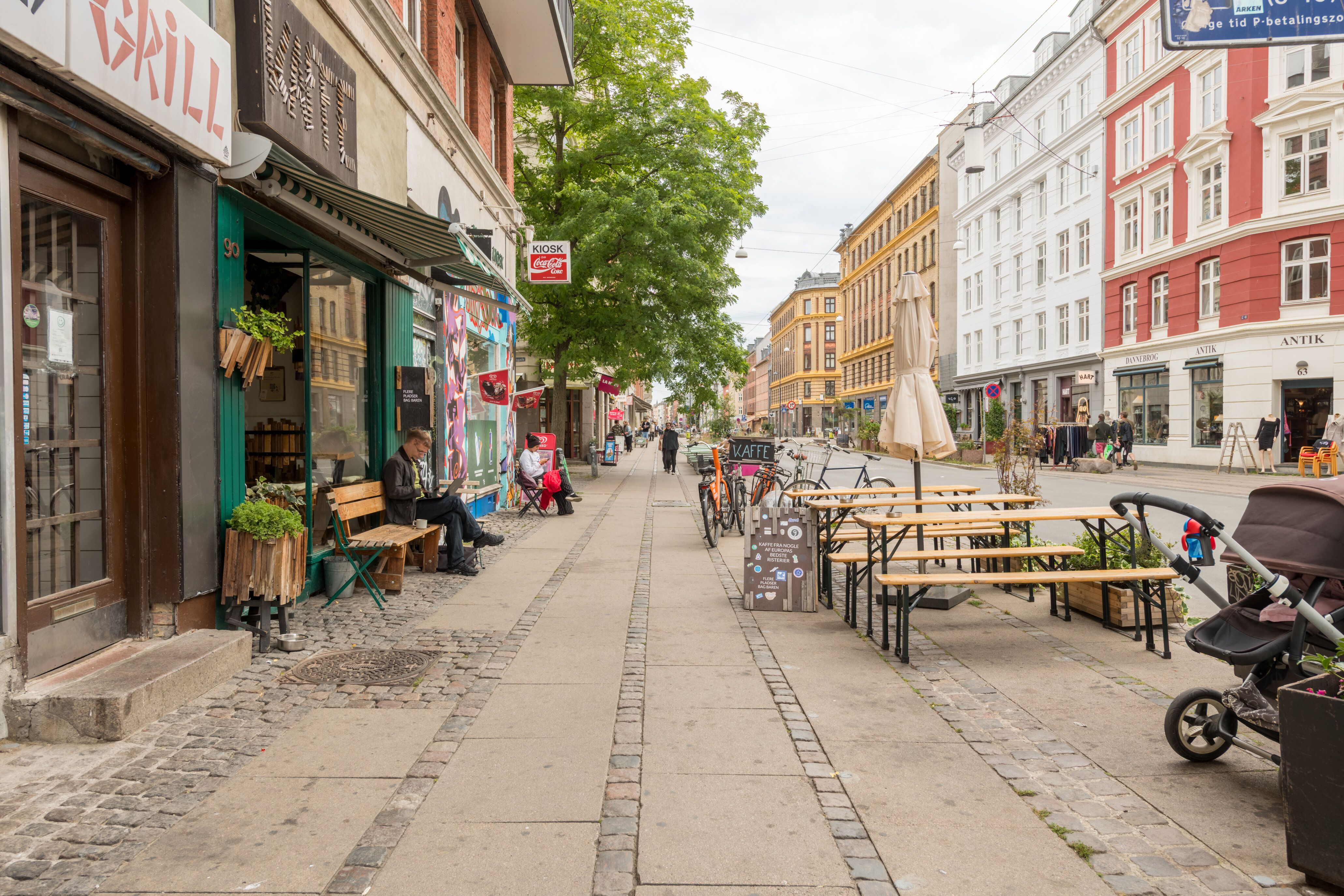 Gade med cafeer og butikker i København. En mand sidder på en bænk og læser. Mange mennesker går rundt. Solskiltet står på en stol.