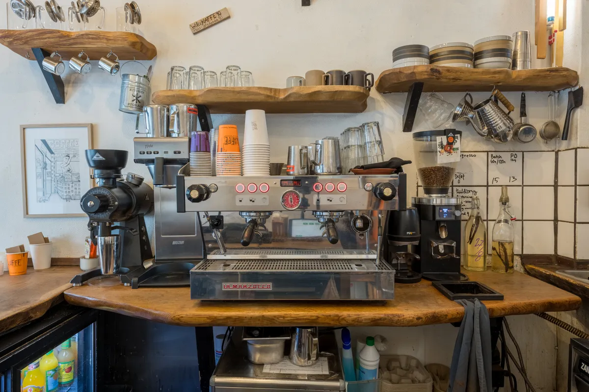 Køkken med kaffemaskiner, krus, glas og hylder med udstyr og apparater til brygning af kaffe og te, ligesom der er en række forskelige krydderier og væskeflasker
