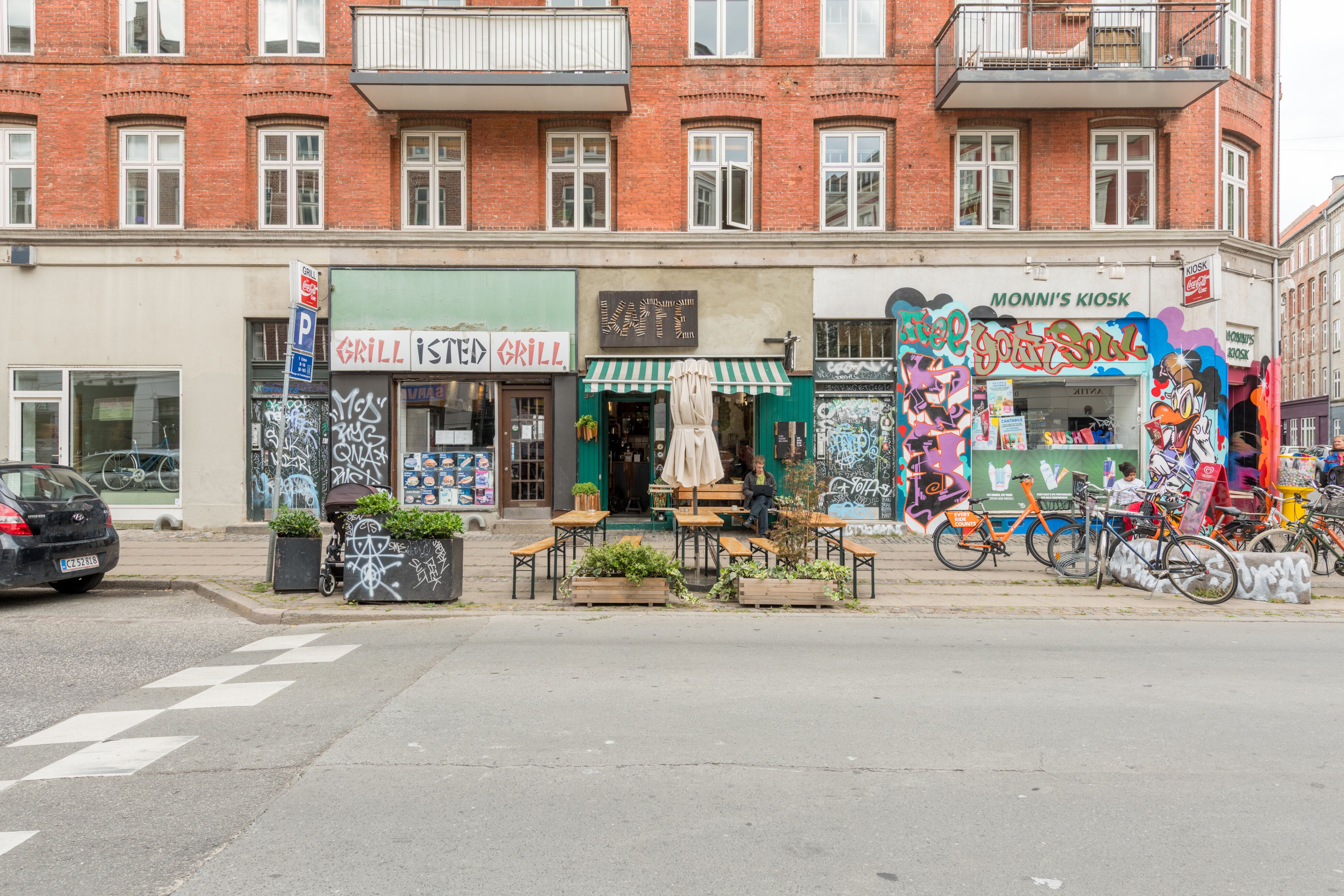 En café med graffiti, træborde, stole, parasolle og planter udenfor, med cykler og skrift på facaden og en bil parkeret på gadens hjørne
