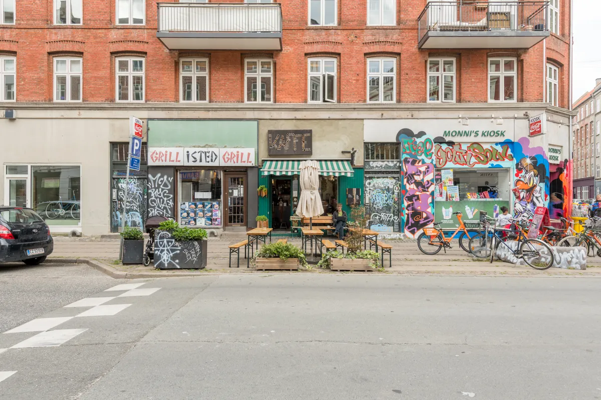 En café med graffiti, træborde, stole, parasolle og planter udenfor, med cykler og skrift på facaden og en bil parkeret på gadens hjørne