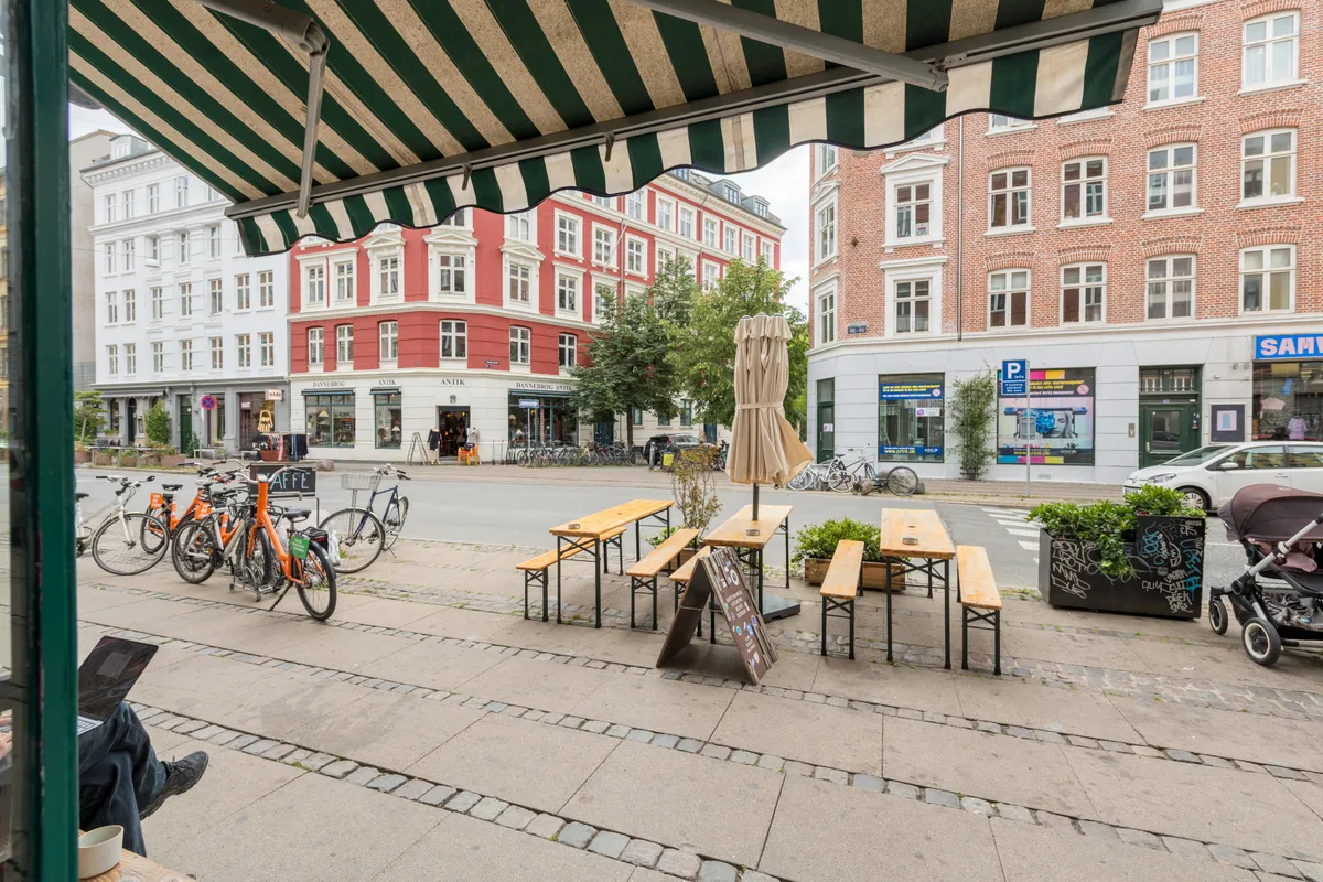 En kafé med udendørsservering og cykelparkering. Parkerede cykler og spisesteder på gågaden. En person sidder med en bærbar computer og en vogn med en baby på gågaden
