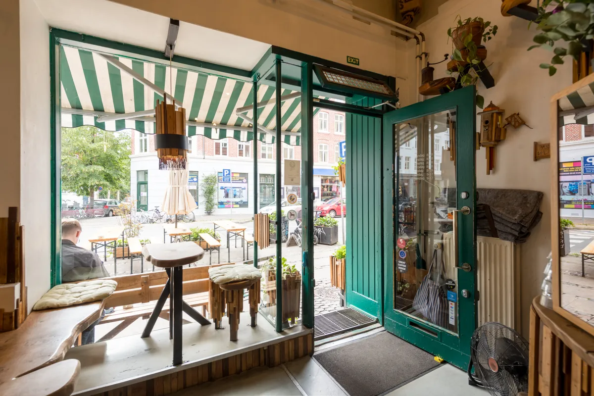En køkkenbord og stole i en café med grønne døre og træstole, plantepot og en lampe, udendørsborde og træbænke, samt en kunde, der sidder og læser