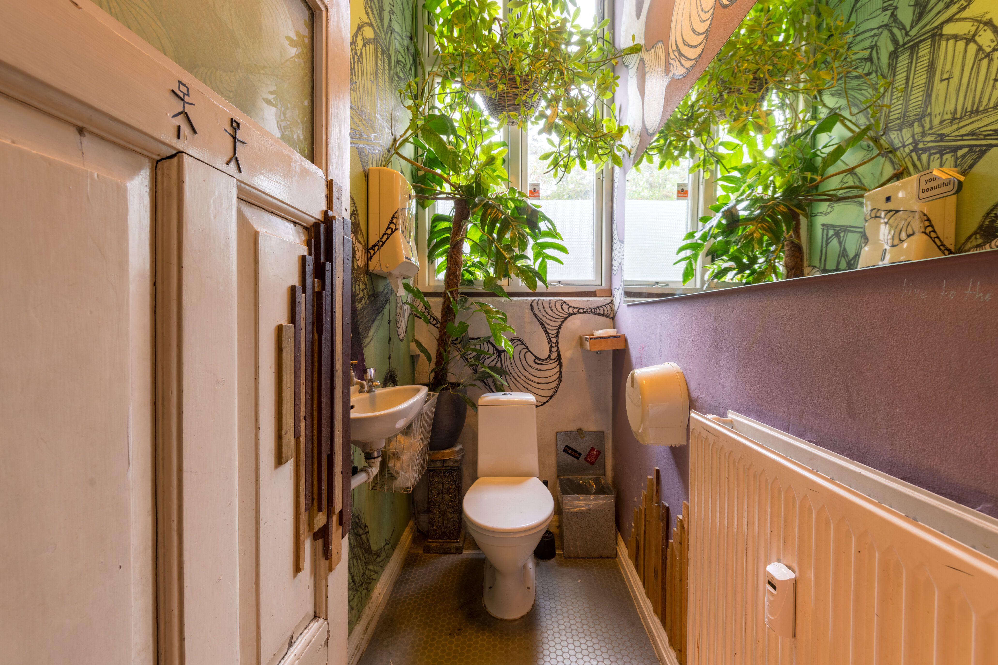Lille toiletrum med toilet, håndvask og toiletpapir med tegninger af mennesker og planter på væggene og loftet.