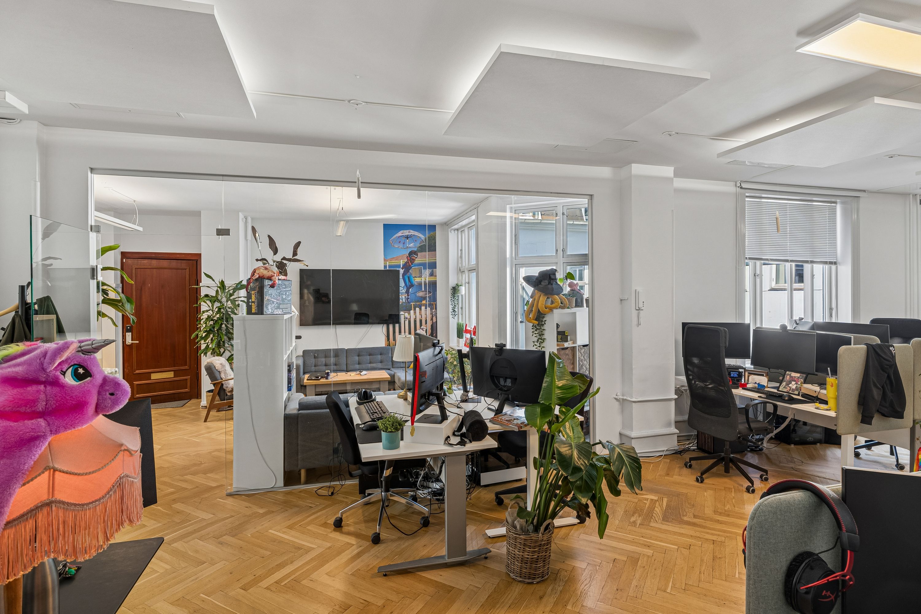Lyst og højloftet kontor med lyse lofter og store ruder, computere, skriveborde og en sofa i baggrunden. Der er flere sjove figurindslag og plantepoter med grønne planter.