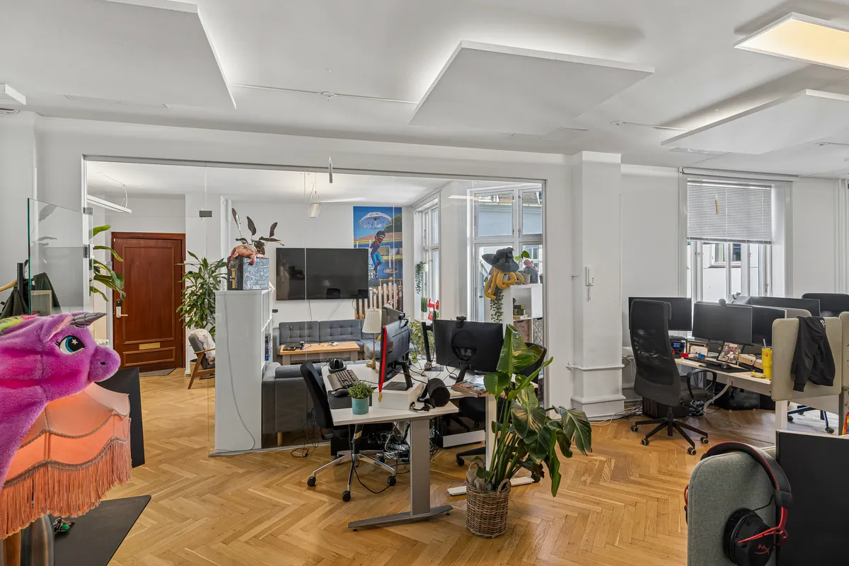 Lyst og højloftet kontor med lyse lofter og store ruder, computere, skriveborde og en sofa i baggrunden. Der er flere sjove figurindslag og plantepoter med grønne planter.