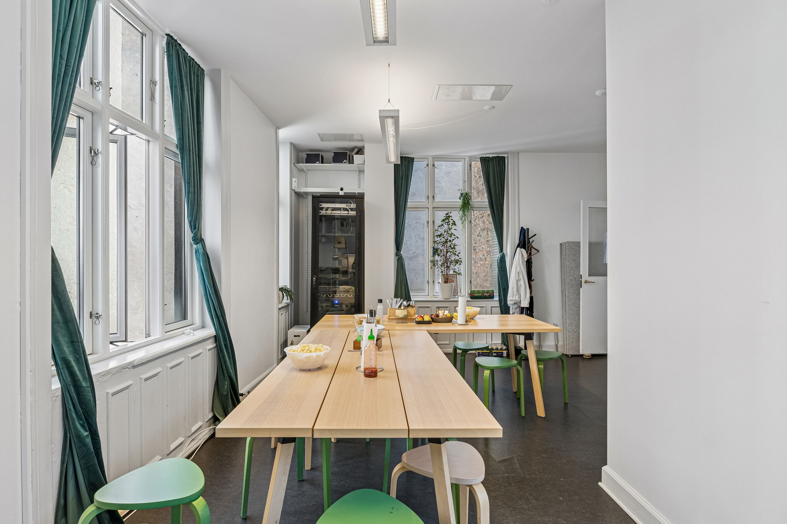 Lyst kontor med lange træborde og grønne stole på frederiksberggade 28, 1459 København K. Vinduer med grønne gardiner og plante på vindueskarmen.
