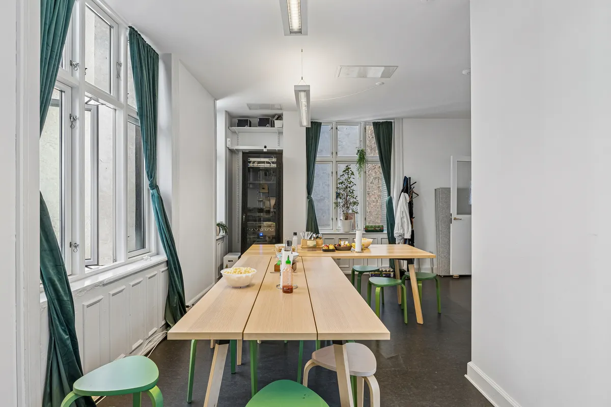 Lyst kontor med lange træborde og grønne stole på frederiksberggade 28, 1459 København K. Vinduer med grønne gardiner og plante på vindueskarmen.