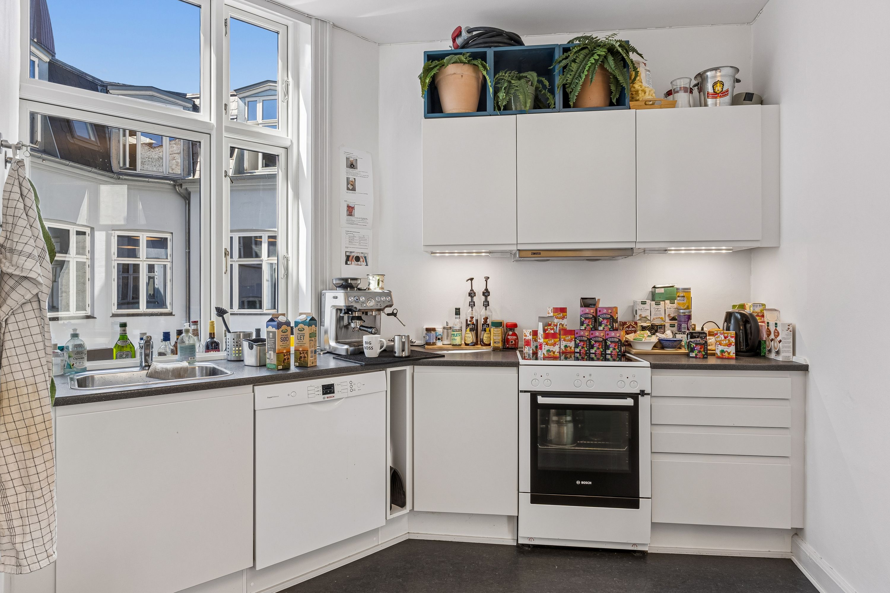 Moderne køkken med lyst vindue, hvidt køkken med Bosch ovn, lyseskabe og møbler samt køkkenredskaber og sager placeret på hylder og bord.