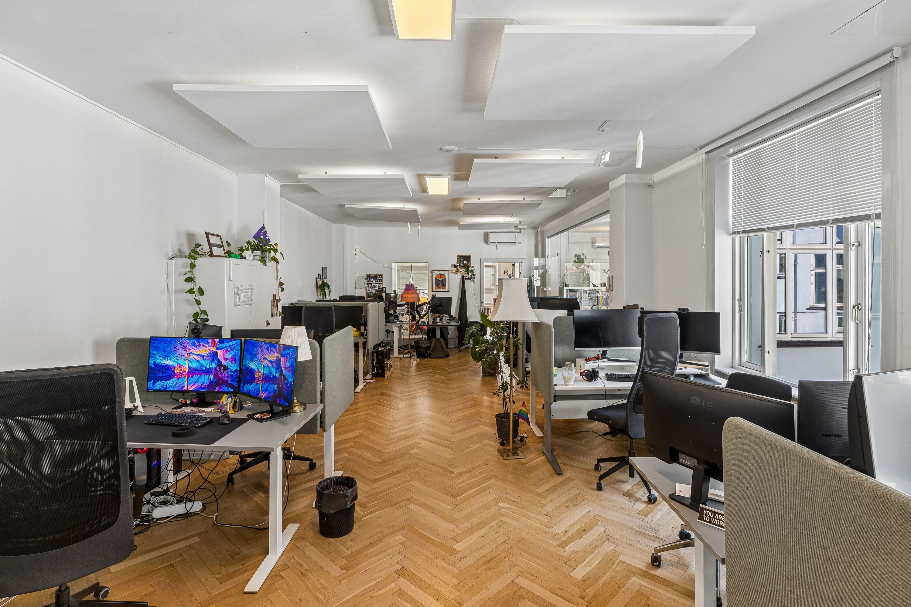 Et moderne kontor med hvide vægge og lyse lofter med lysekroner. Arbejdsborde med computere, planter og flere stole er opstillet i et slankt design. Et slankt design med lyse vægge og trægulve.