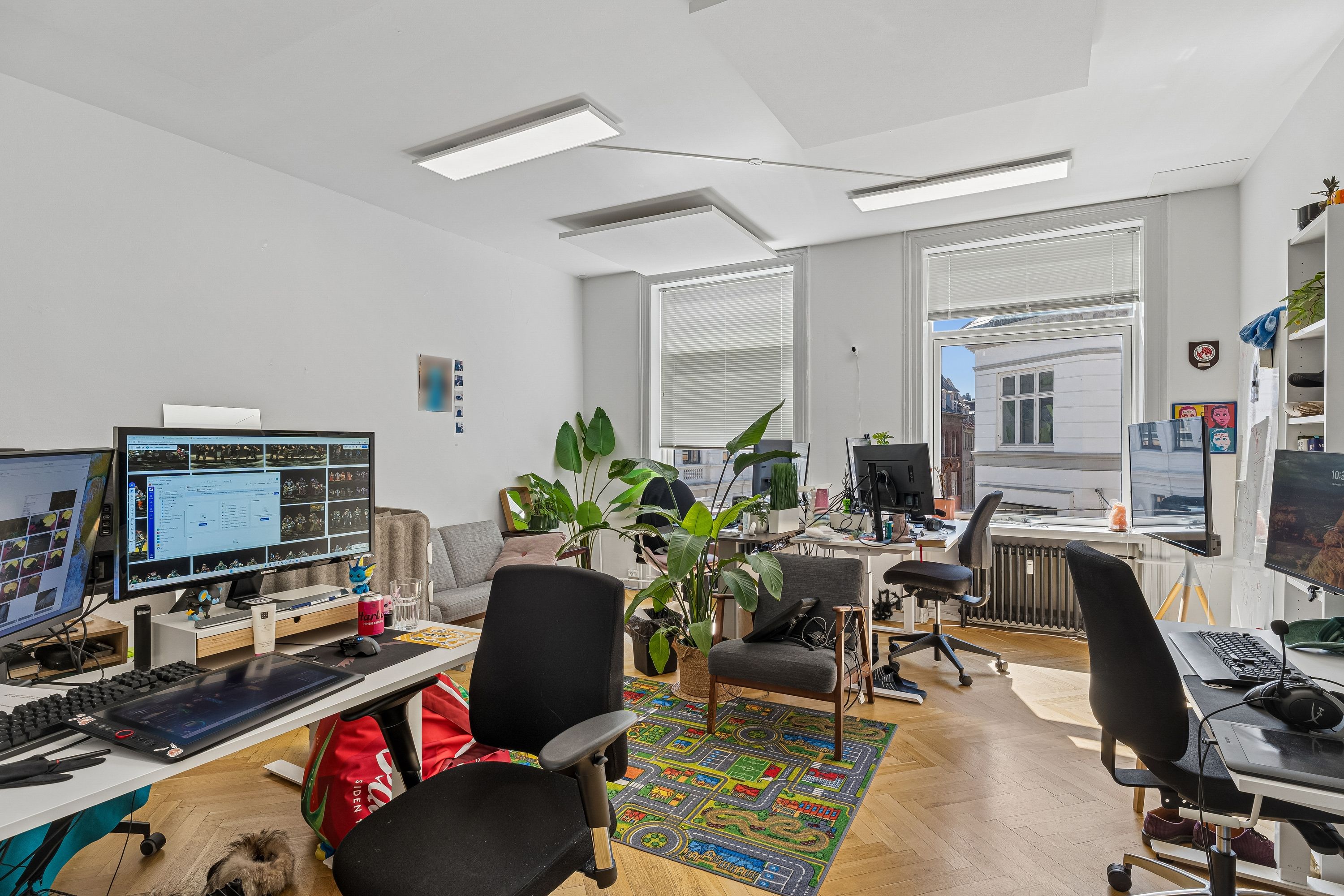 Et kontor med mange skriveborde, computere og plante. Bord med tastatur og tavle, sofa og vinduessjalusier. Moderne møbler og lyse lofter.