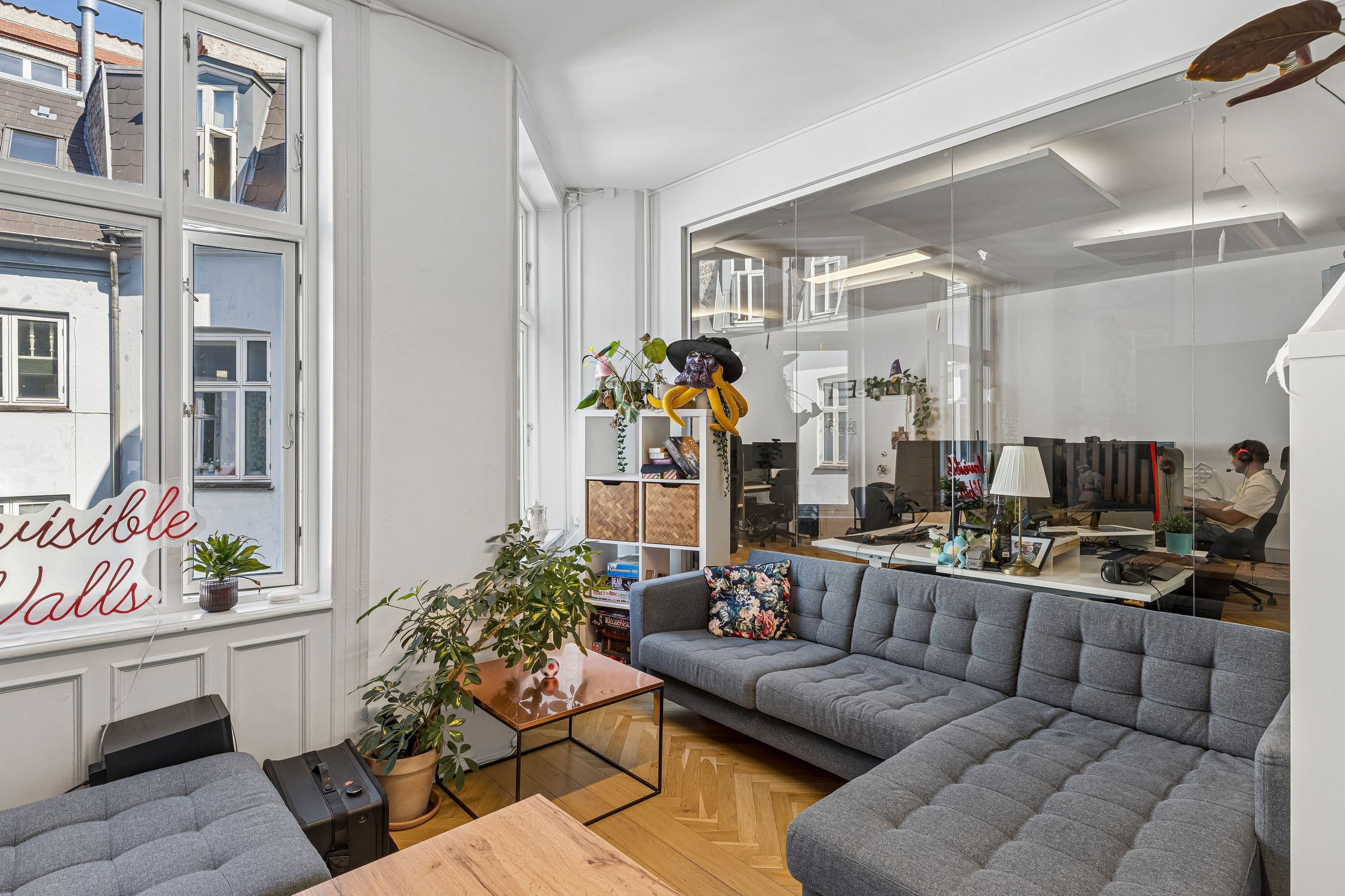 Kontor til leje på Frederiksberggade med lyse vinduer og smukke glasvægge. I stuen står sofaen, træbord og en plante. Kontoret er fuldt udstyret med computere og kontormøbler.