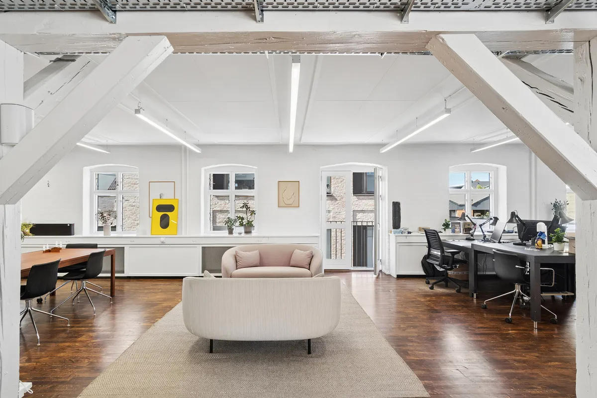 Moderne kontor med lyse loftsbjælker og trægulv, skriveborde og stole i minimalistisk stil og et kunstværk på væggen