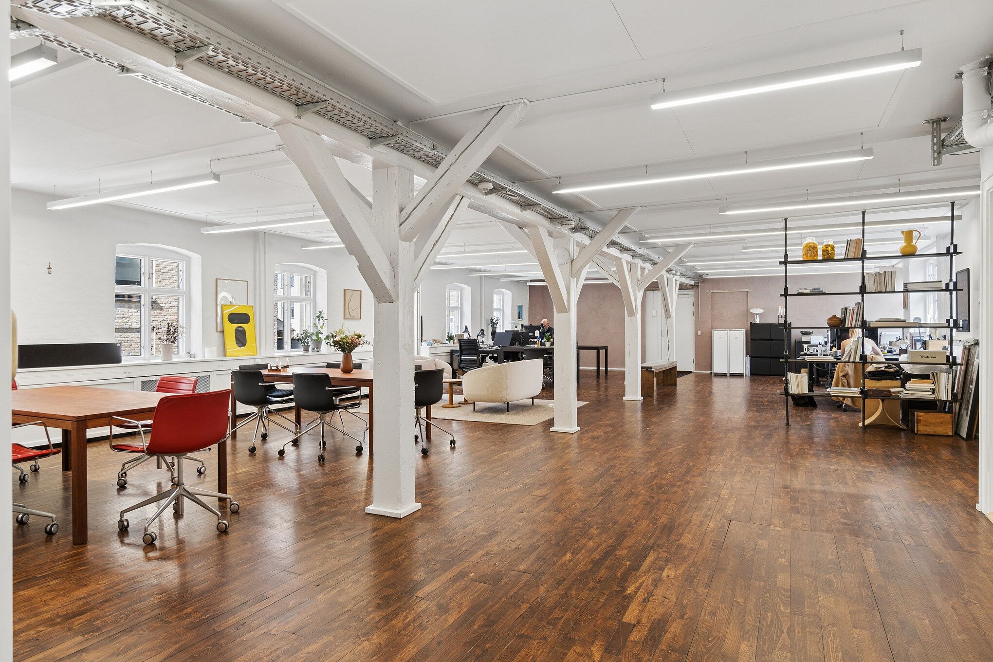 Minimalistisk kontor med højt loft, hvide bjælker og lyse loftslampe. Bord, stole og sofaer samt bibliotek og hylder med lyse farver og genstande.