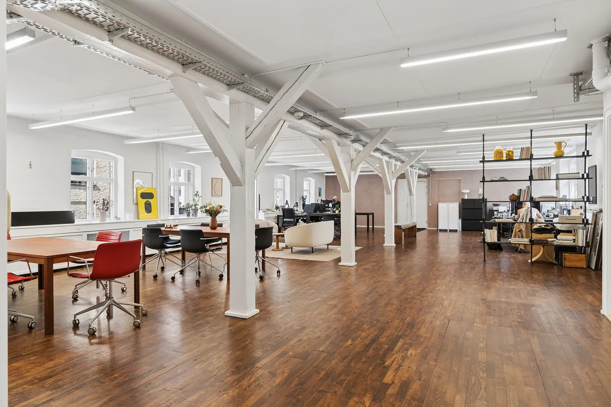 Minimalistisk kontor med højt loft, hvide bjælker og lyse loftslampe. Bord, stole og sofaer samt bibliotek og hylder med lyse farver og genstande.