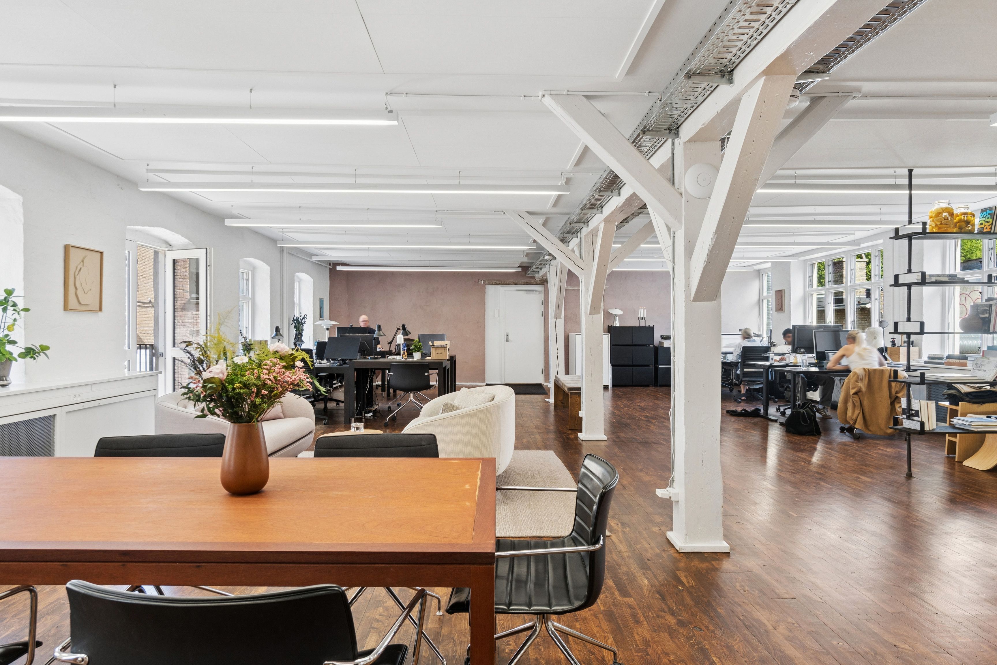 Et moderne kontor med højt til loftet og lyse loftsbjælker samt gult gulv og hvide vægge. Kontoret har stole, en stor bord, blomster og et plante på vindueskarmen