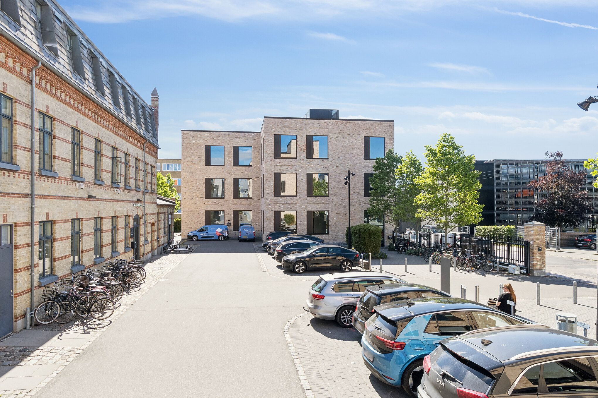 Moderne kontorbygning med glasvinduer og røde mursten. Parkerede biler og cykler samt træer og buske langs vejen.