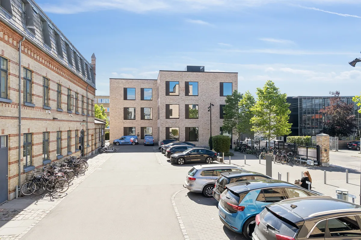 Moderne kontorbygning med glasvinduer og røde mursten. Parkerede biler og cykler samt træer og buske langs vejen.