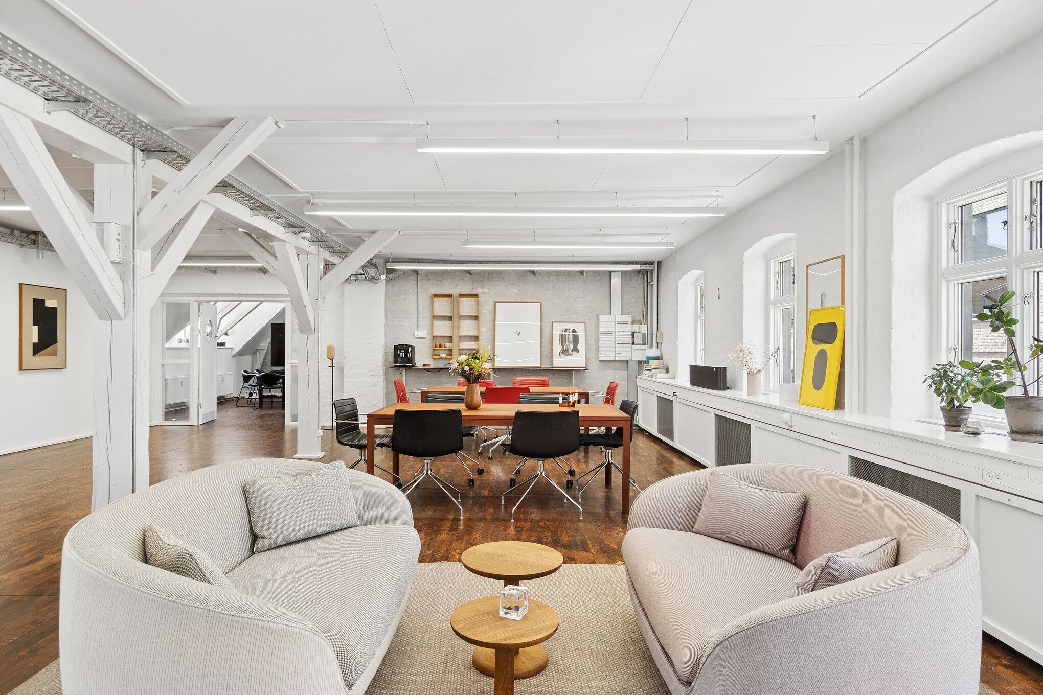 Modernt kontor med højt loft, lyse loftsbjælker, to buede højryggede stole og massivt træbord med røde stole. Hjørne med blomster og lyse farver på væggen og lyse gulve