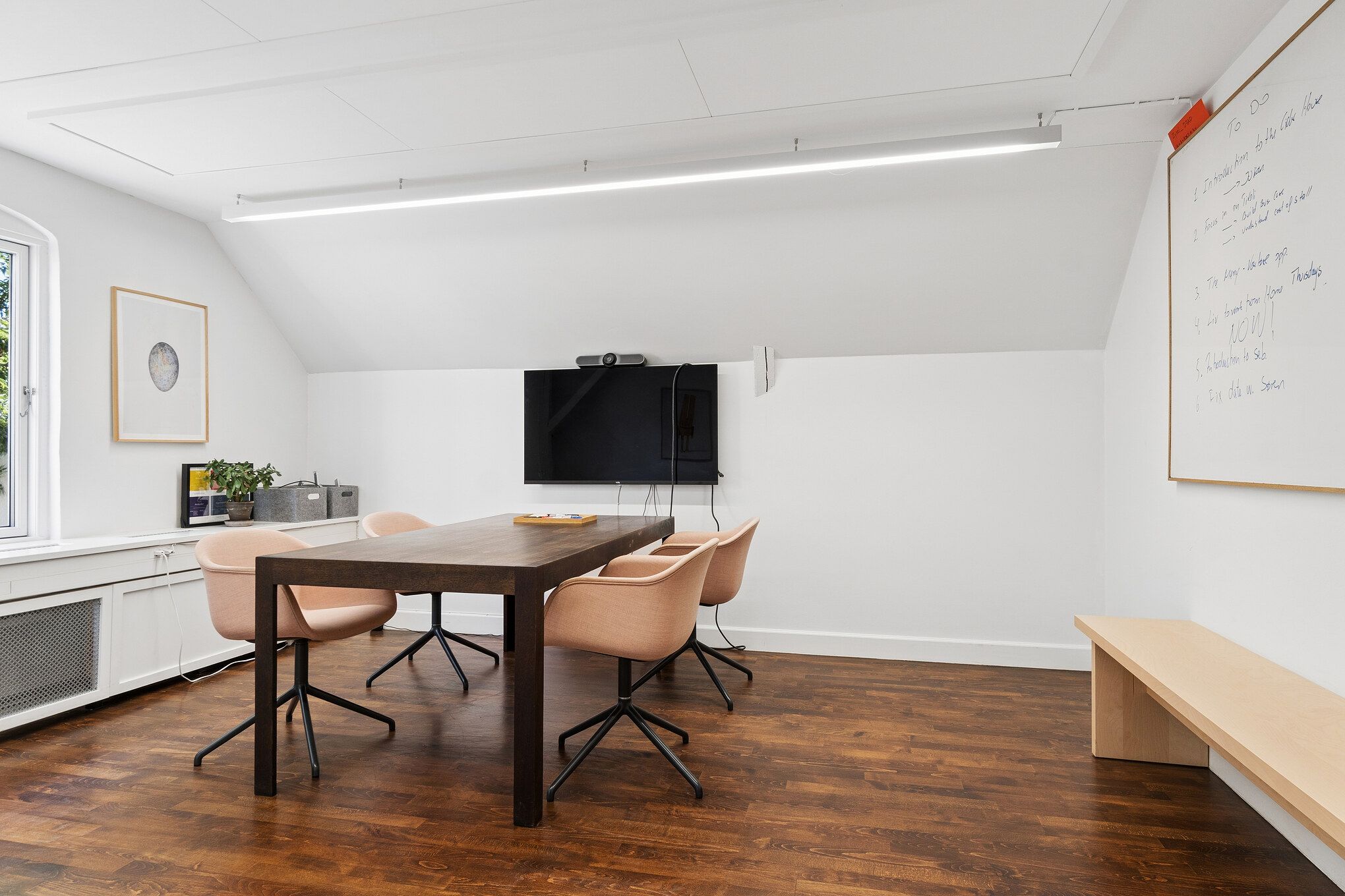 Minimalistisk kontor med lysgrå vægge og trægulv, bord og stole, tv-skærm og whiteboard i hvidt, møbler i lyse nuancer, en plante og maleri i hjørnet