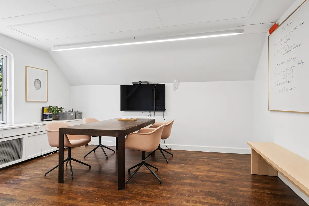 Minimalistisk kontor med lysgrå vægge og trægulv, bord og stole, tv-skærm og whiteboard i hvidt, møbler i lyse nuancer, en plante og maleri i hjørnet
