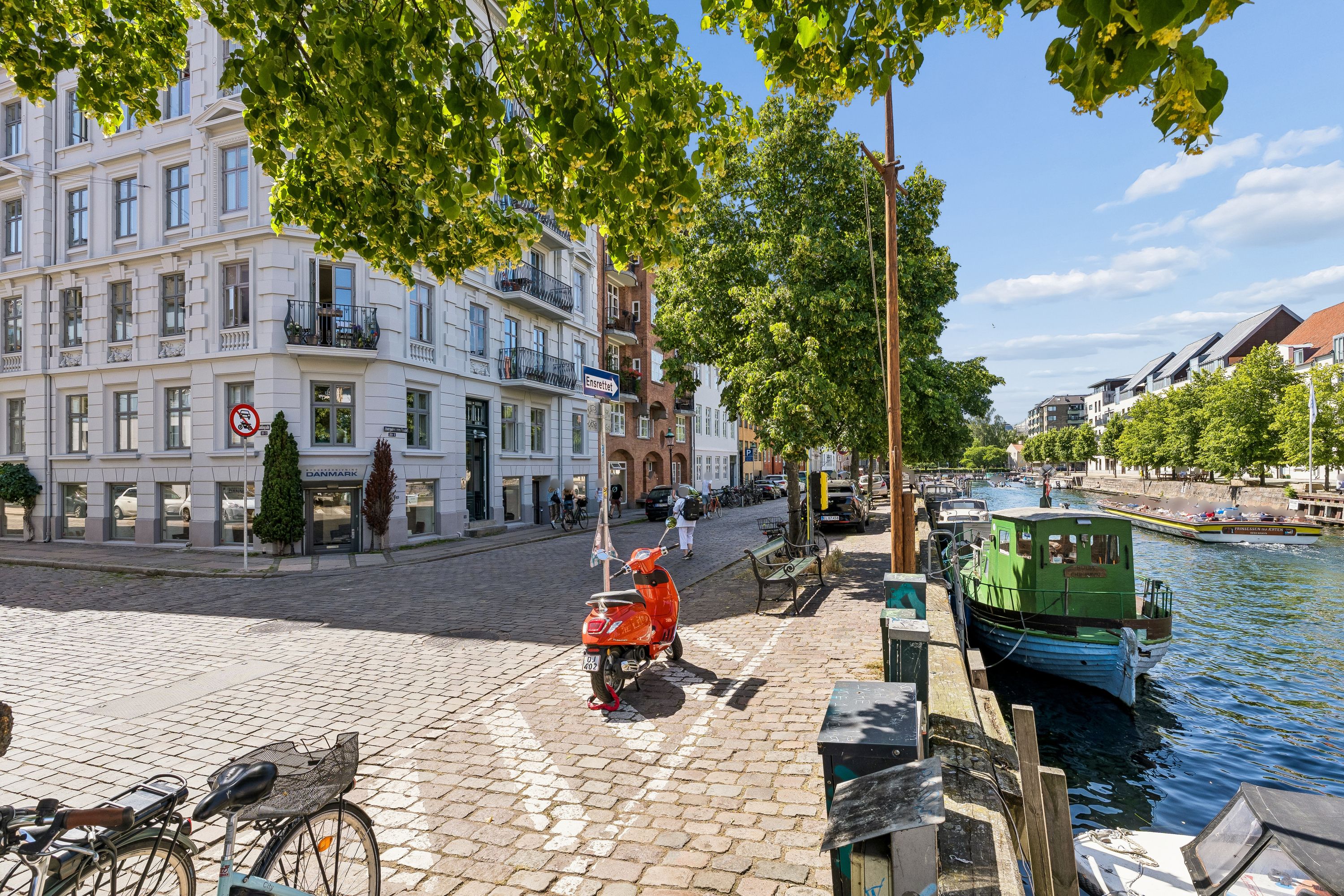 Københavns kanal med ro-ro-båd, motorcykel og cykler parkeret og gående folk på broen og i baggrunden står høje bygninger