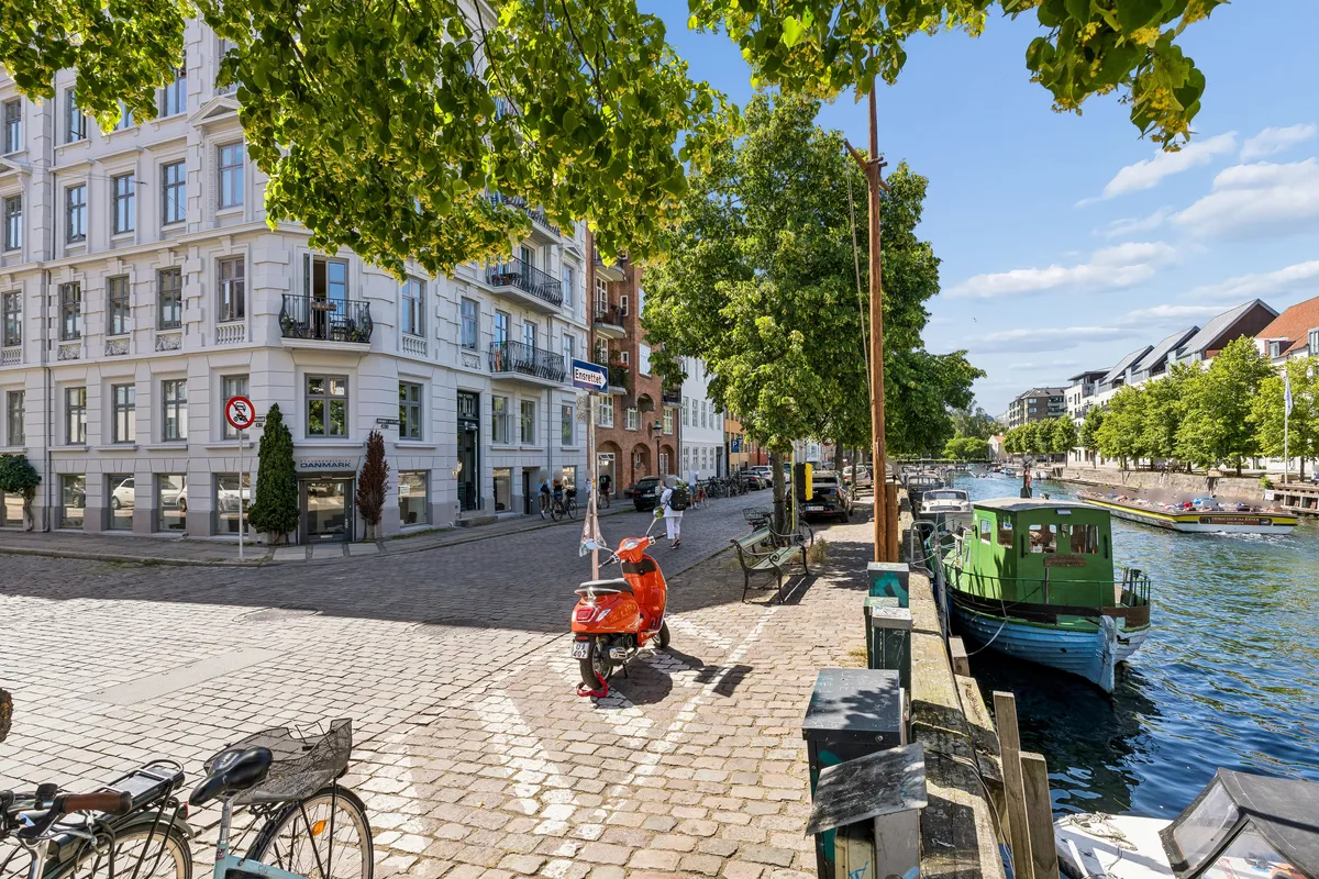 Københavns kanal med ro-ro-båd, motorcykel og cykler parkeret og gående folk på broen og i baggrunden står høje bygninger