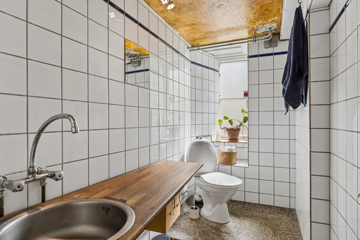 En lille wc-rum med en trægulv og hvide flisede vægge. Et toilet står i rummet, og et håndtag er fastgjort til væggen. En plante står i vindueskarmen, og et håndklæde hænger på væggen.