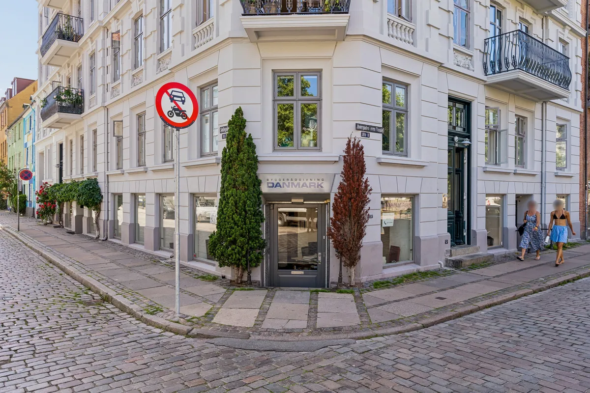 Kontor på Overgaden Oven Vandet 30B med indgangsdør, facade med røde planter og en skilt med teksten Danmark. To kvinder går langs fortovet med små tasker