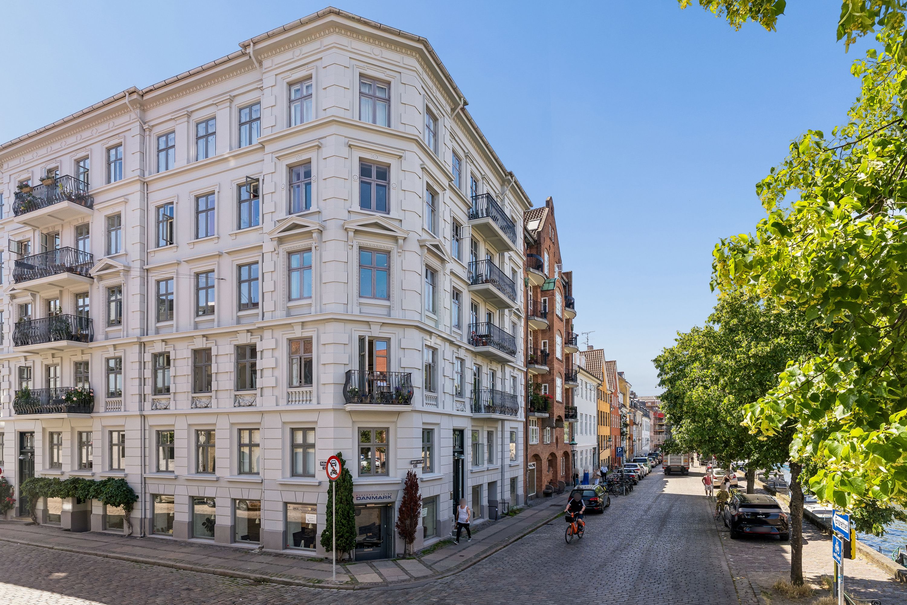Kontor til salg på Overgaden Oven Vandet 30B, København. Bygning med lyse fasadebelægning og store vinduer. På gaden cykler en person, og biler er parkeret langs gaden.