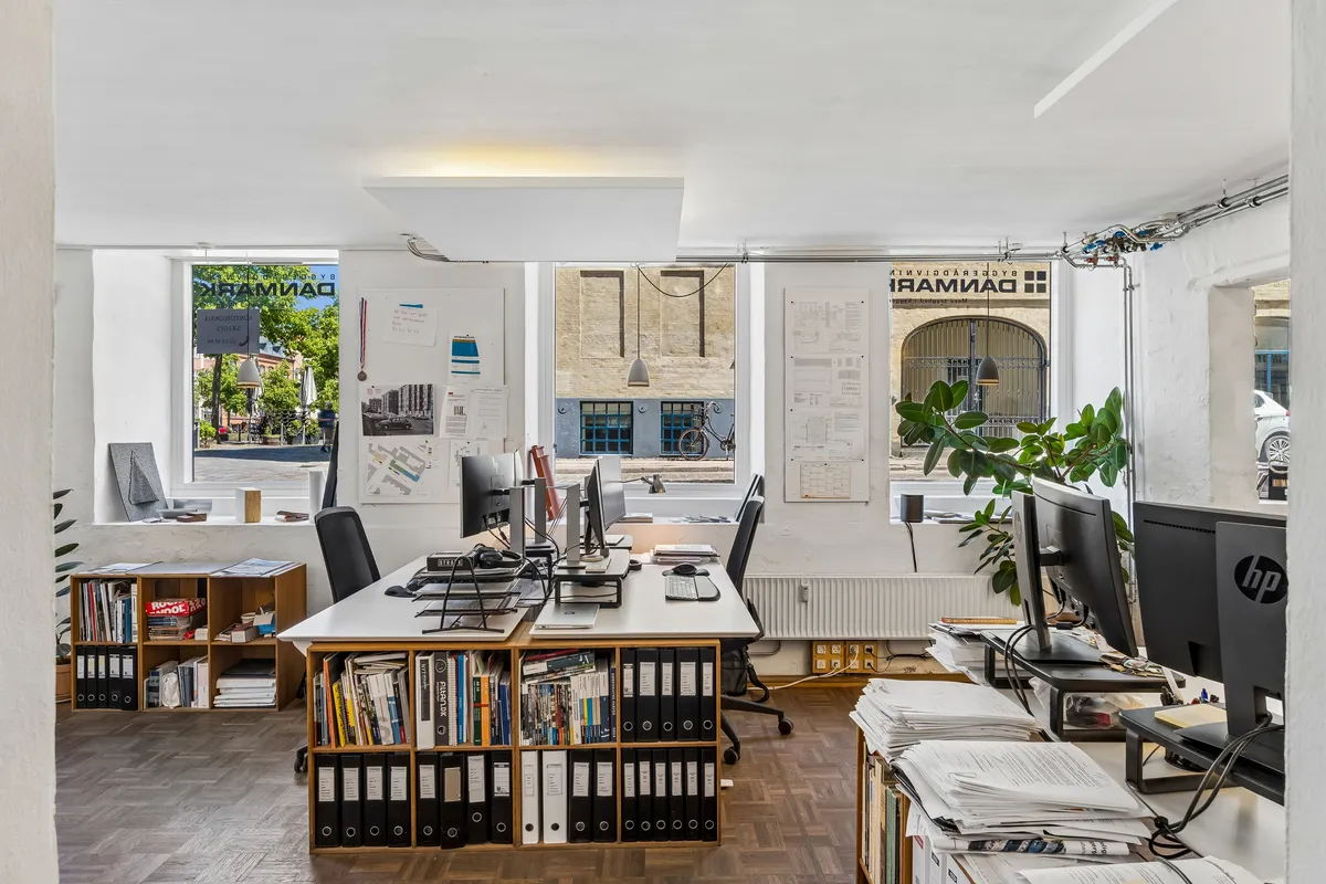 Kontor til salg i København med fuld udsigt over gaden og gamle bygninger, store vinduer og lyse lofter, moderne computerudstyr og hyggeligt skrivebord.