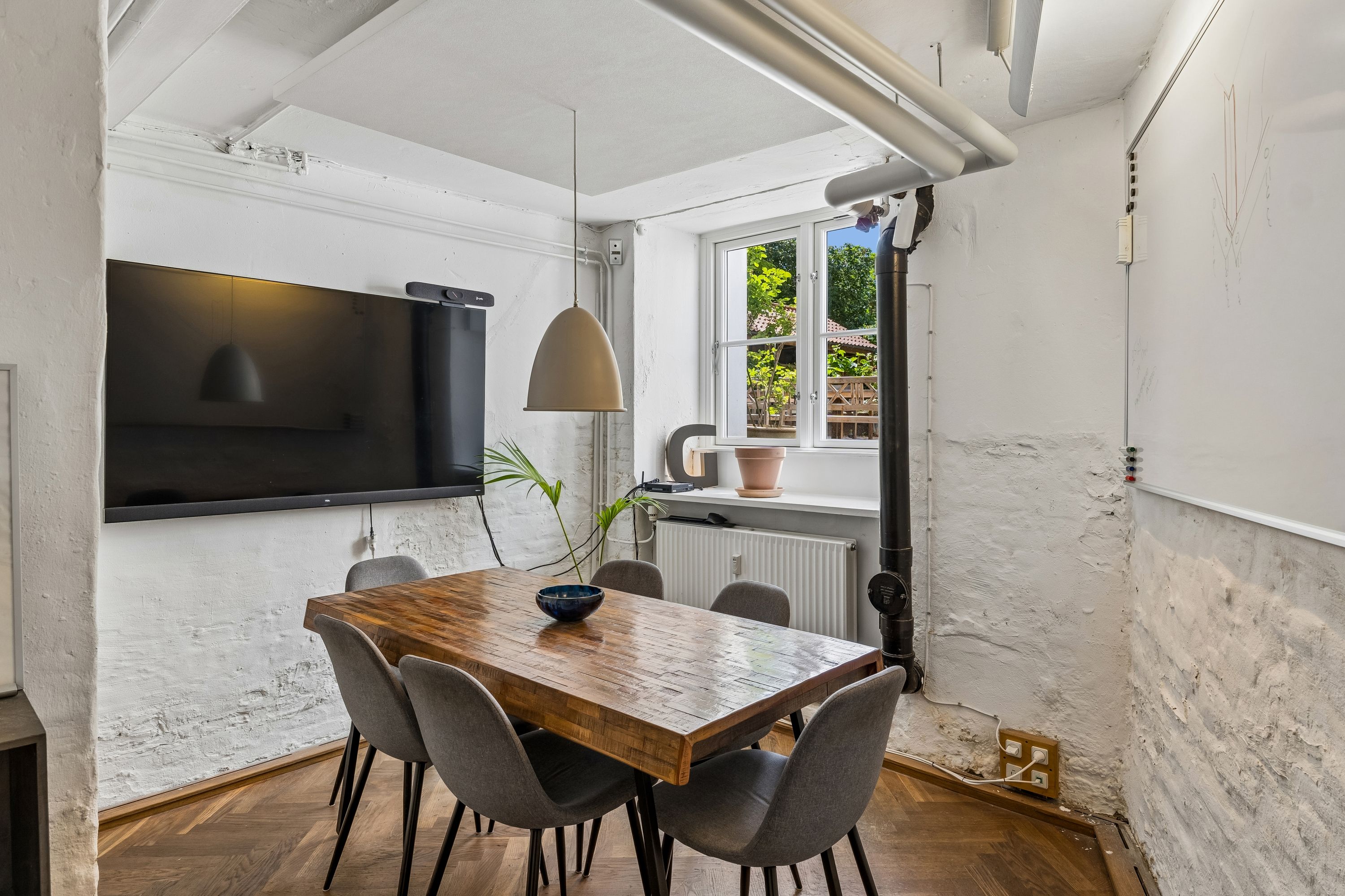 Kontor med moderne møbler, bord og stole, plante og tv på væggen, radiator og vinduer med udsigt til haven