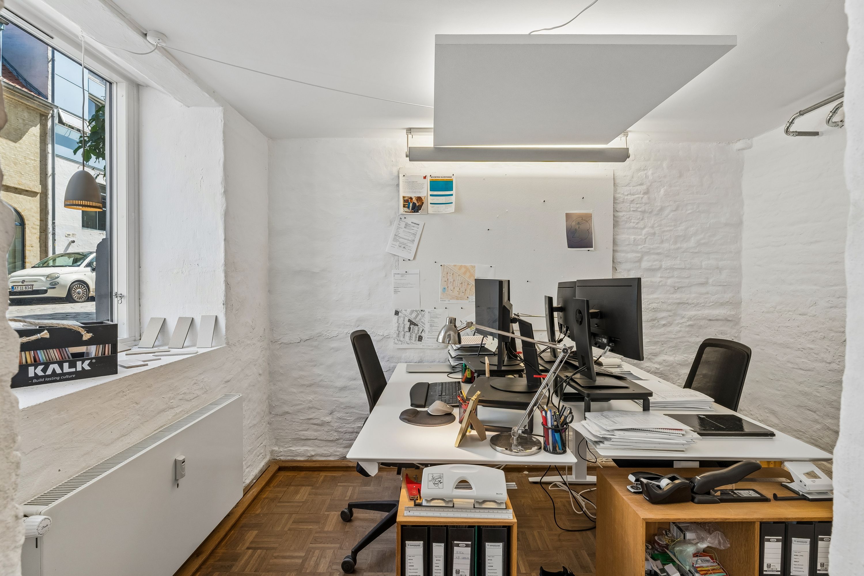 Kontor med skrivebord, computere, lampe, bøger og papir på en høj hylde og kontorvæg med noter
