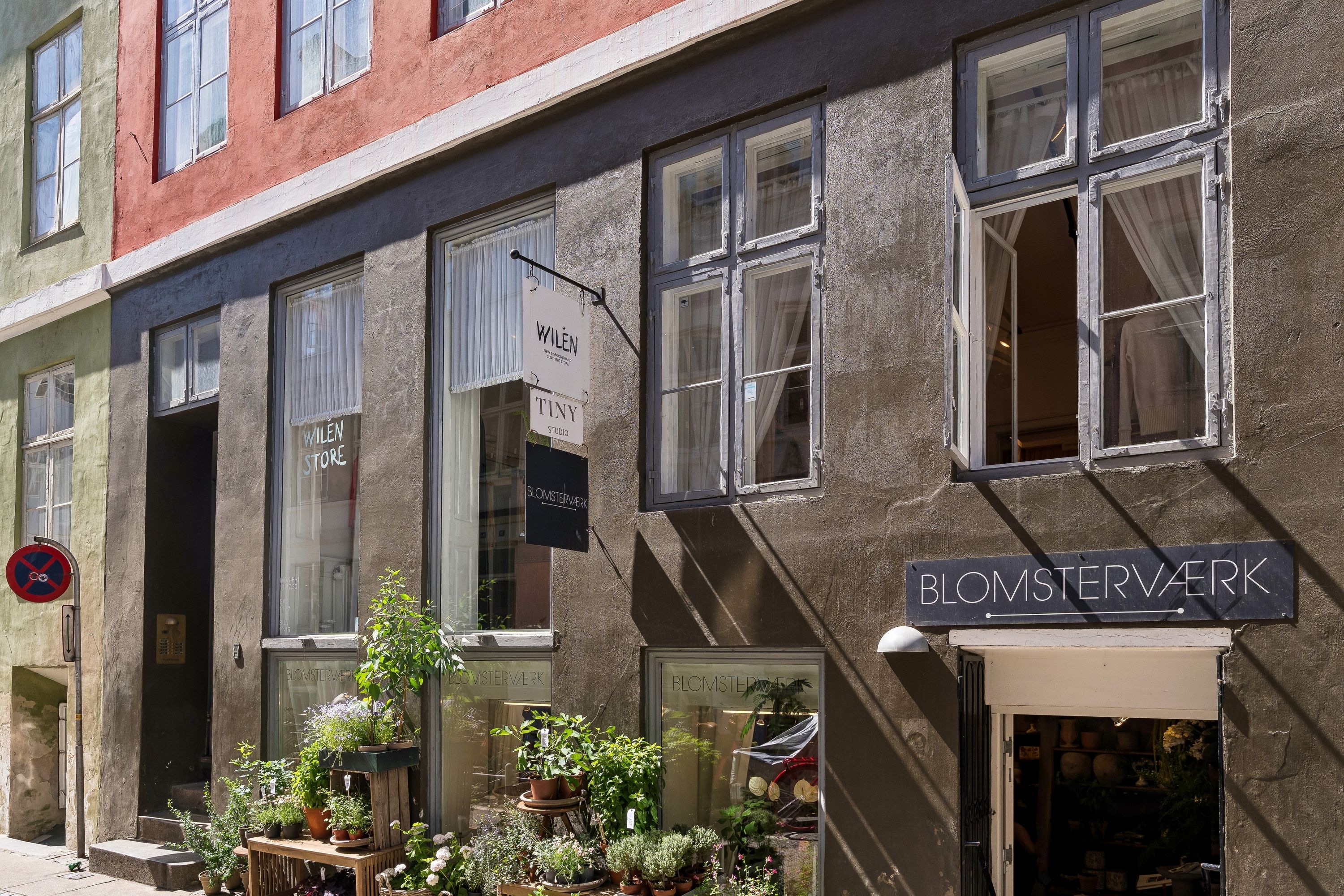 Blomsterværk butik med lyse vinduer og møbler. På gaden er der en række potter med planter og en stol.