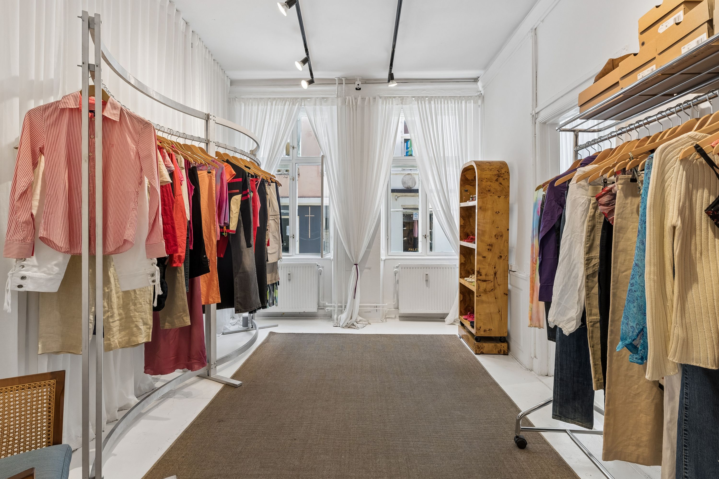 Lys og rummelig butik med klædeskab, hængeklæder, hvidt gulv og lysegardiner. Lyse og farverige tøjstørrelser og -typer. Moderne indretning med lyse og rene vægge