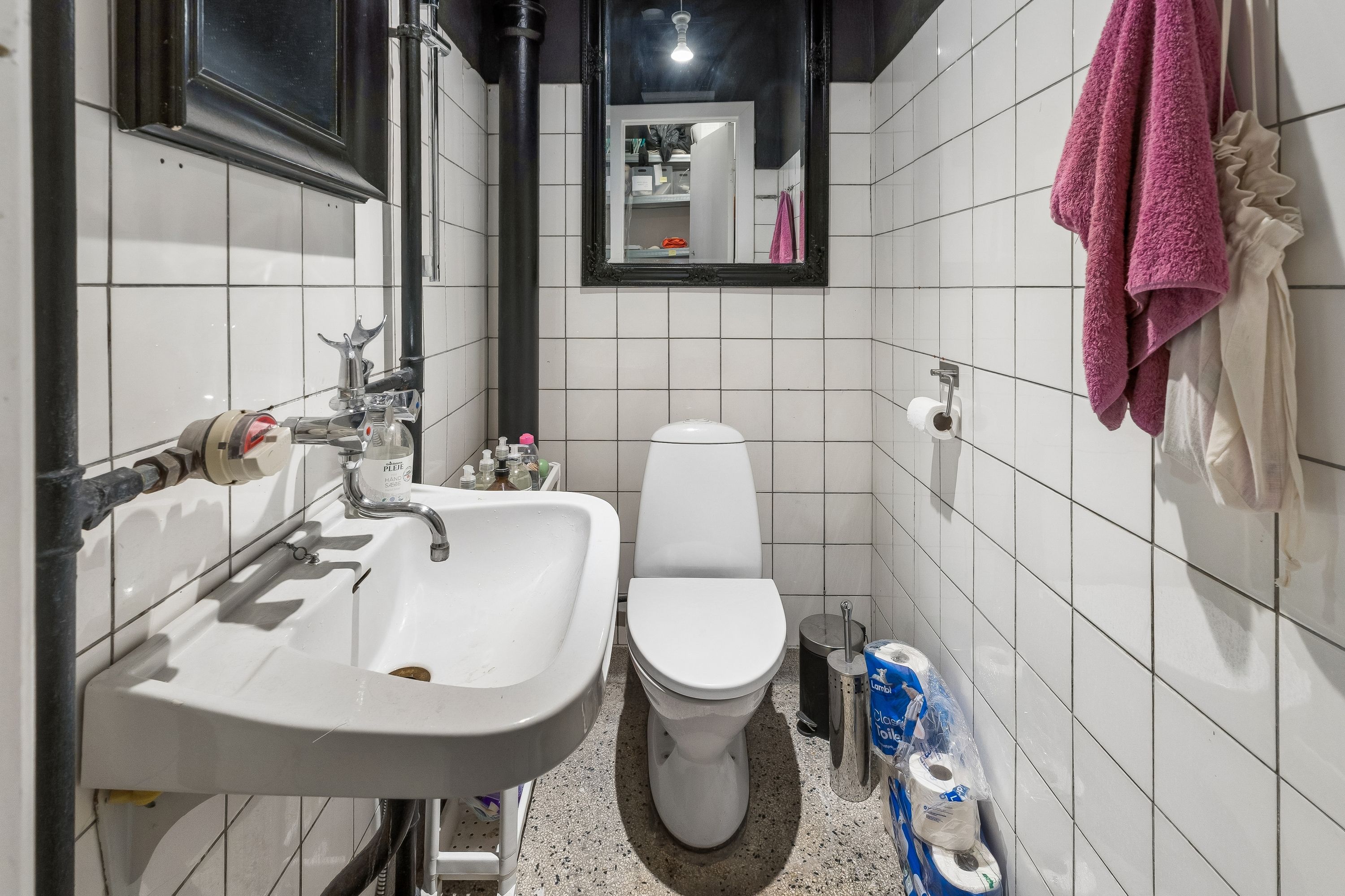 Billet for leje butik i København med toilet, håndvask og tøjvask. Bag toilettet er en række toiletpapir, og en pink håndklæde er hængende på væggen.