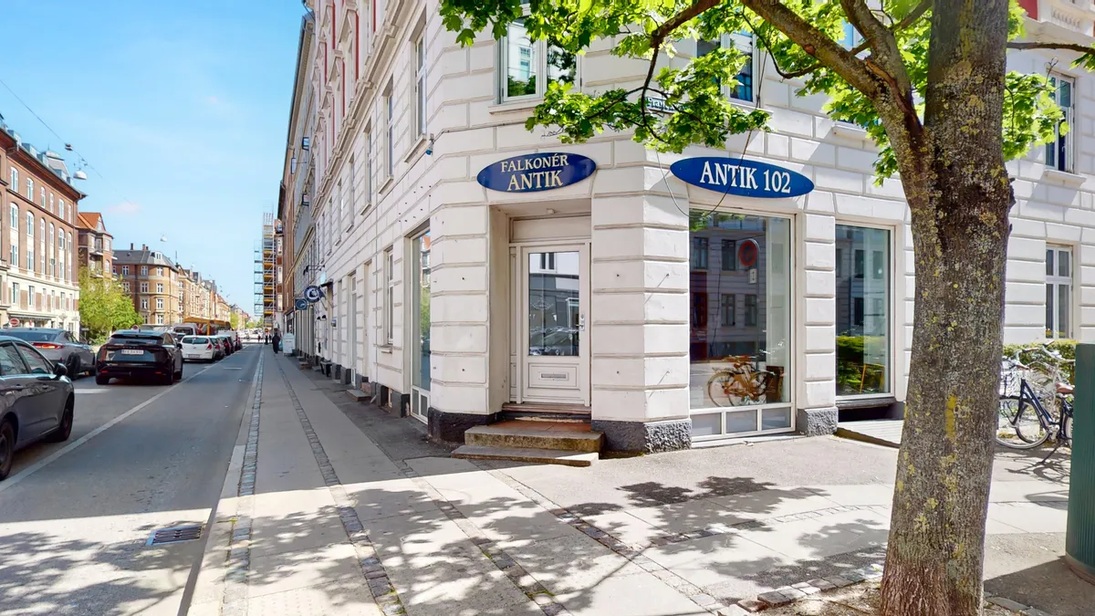 Frederiksberg bymidte med antik butik og gågade. Biler parkeret på siden af gaden. Træ og grønne planter udenfor butikken. Solskin og skygge på gaden.