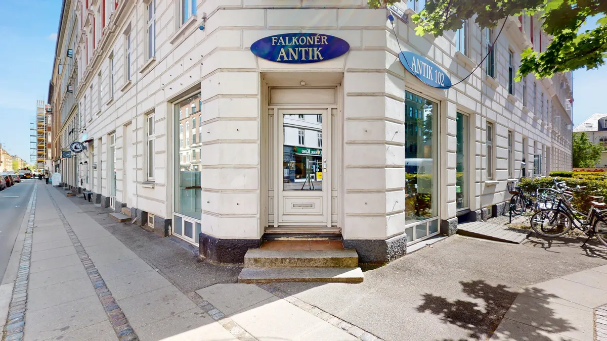 En butik med vinduer og hvide vægge med to schilde med teksten Falckoner Antik og Antik 102. Adgangsdøren er hvid og er åben, og der er cykler parkeret på forsiden