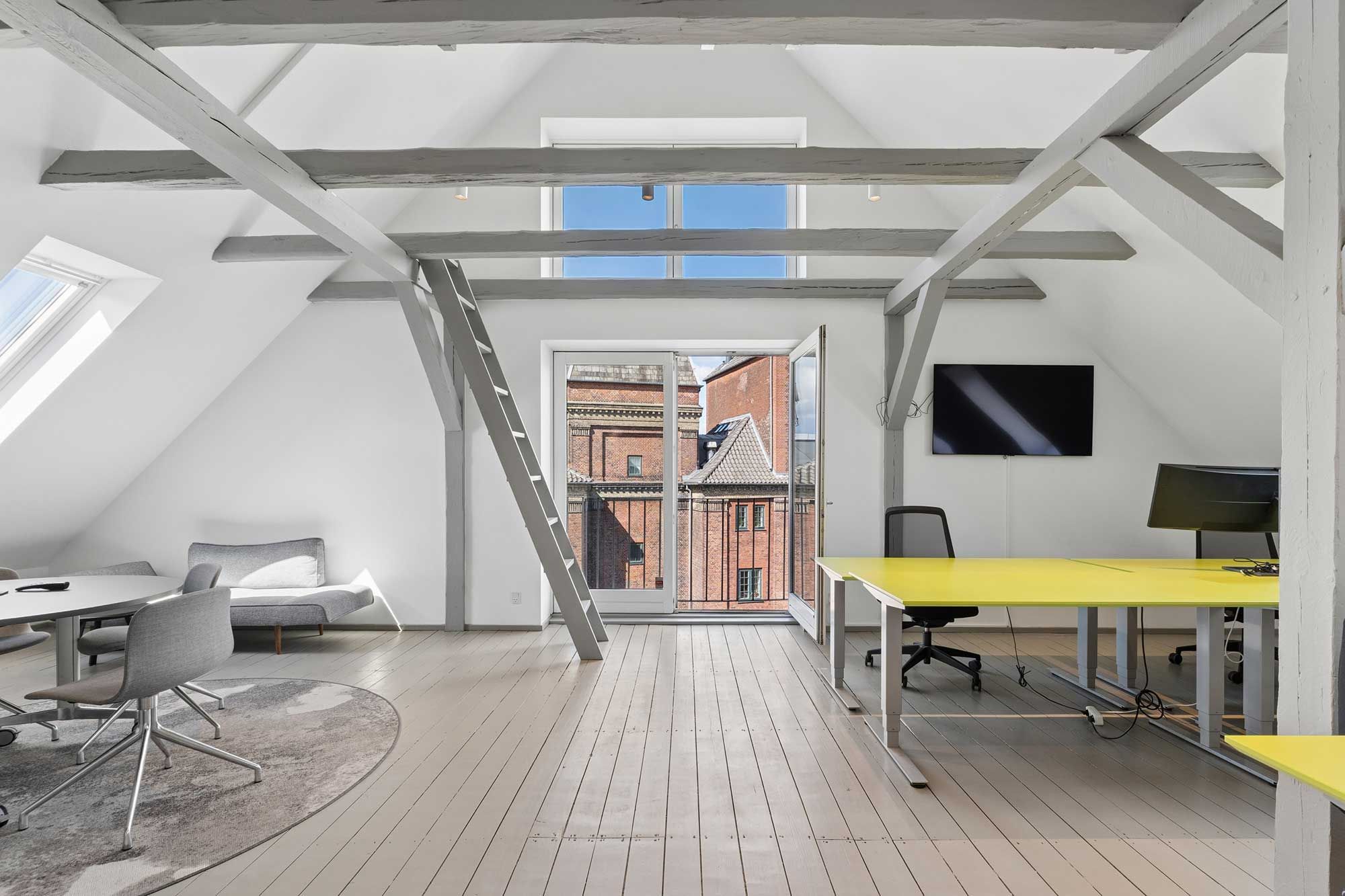 Loftsrummet er indrettet med lyse vægge og lyse træbjælker, der giver en følelse af åbenhed og komfort. To runde borde og stole med moderne design.