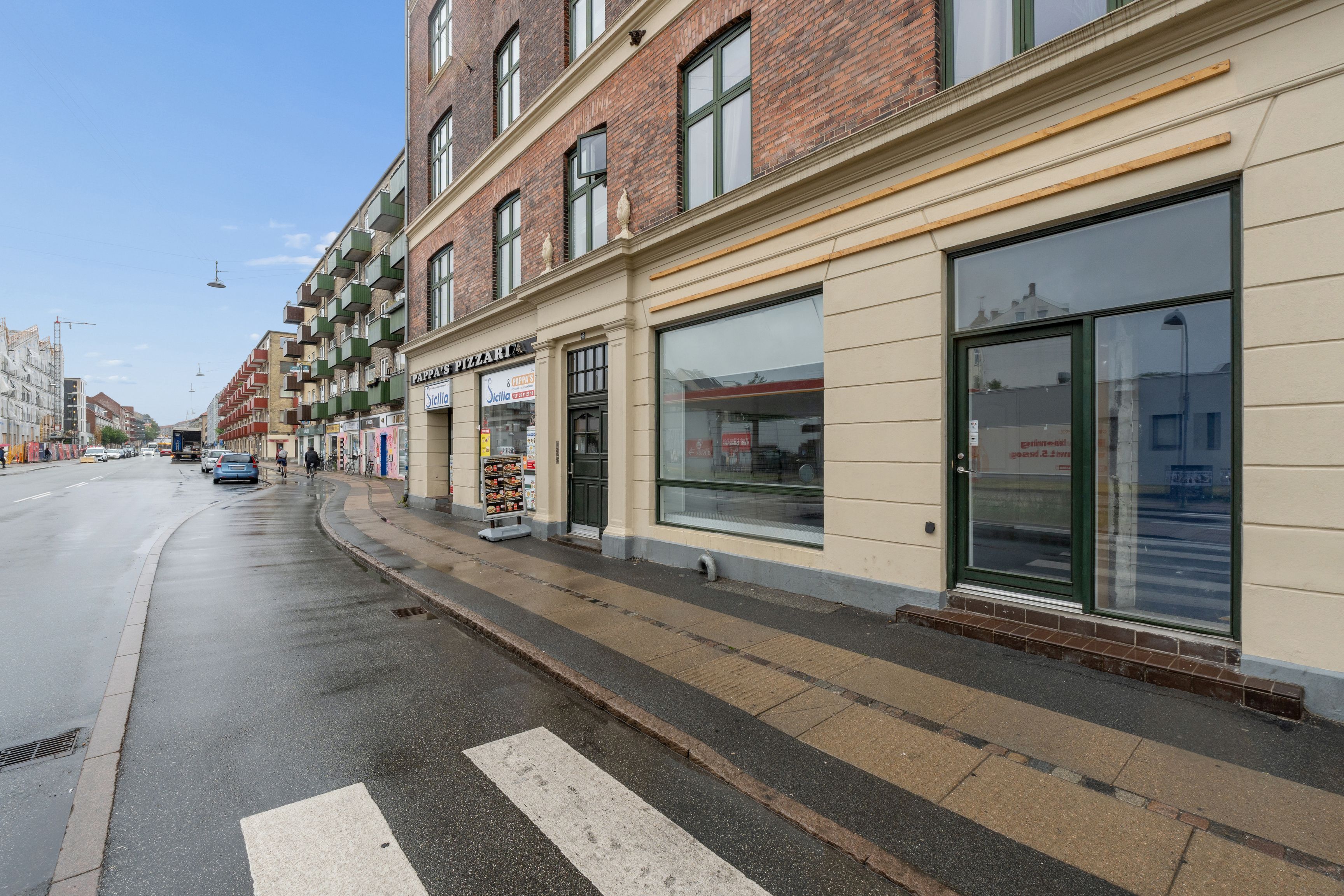 Erhvervslejemål med store glasvinduer og facade i lyse mursten. Lejlighederne er lyse og har gode indskud. Gaden er lige med parkerede biler og gågade.