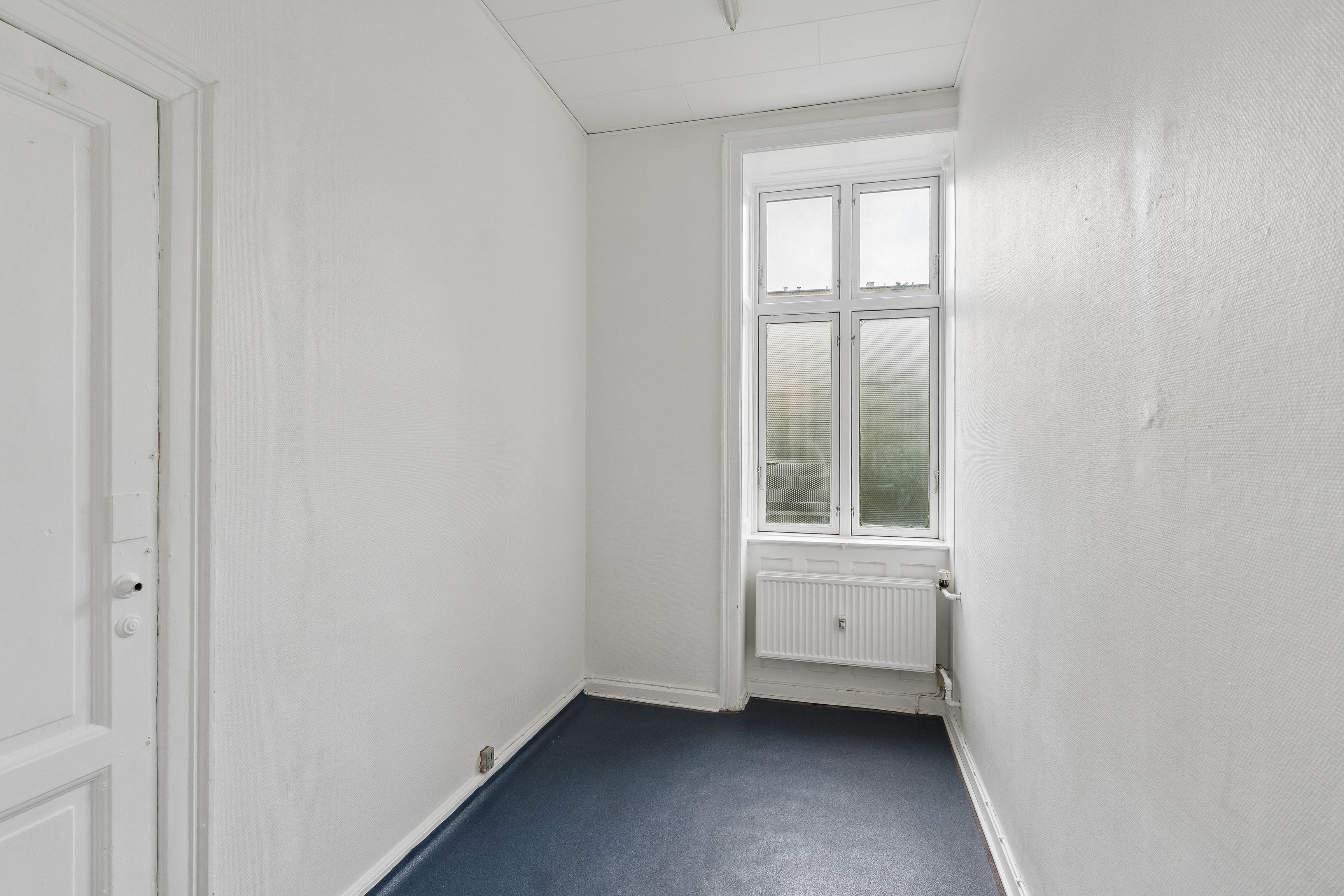 En tom rummelig værelse med hvide vægge og gulvtæppe, et vindue med dobbeltruder og radiator samt en hvid dør med to håndtag og et lås