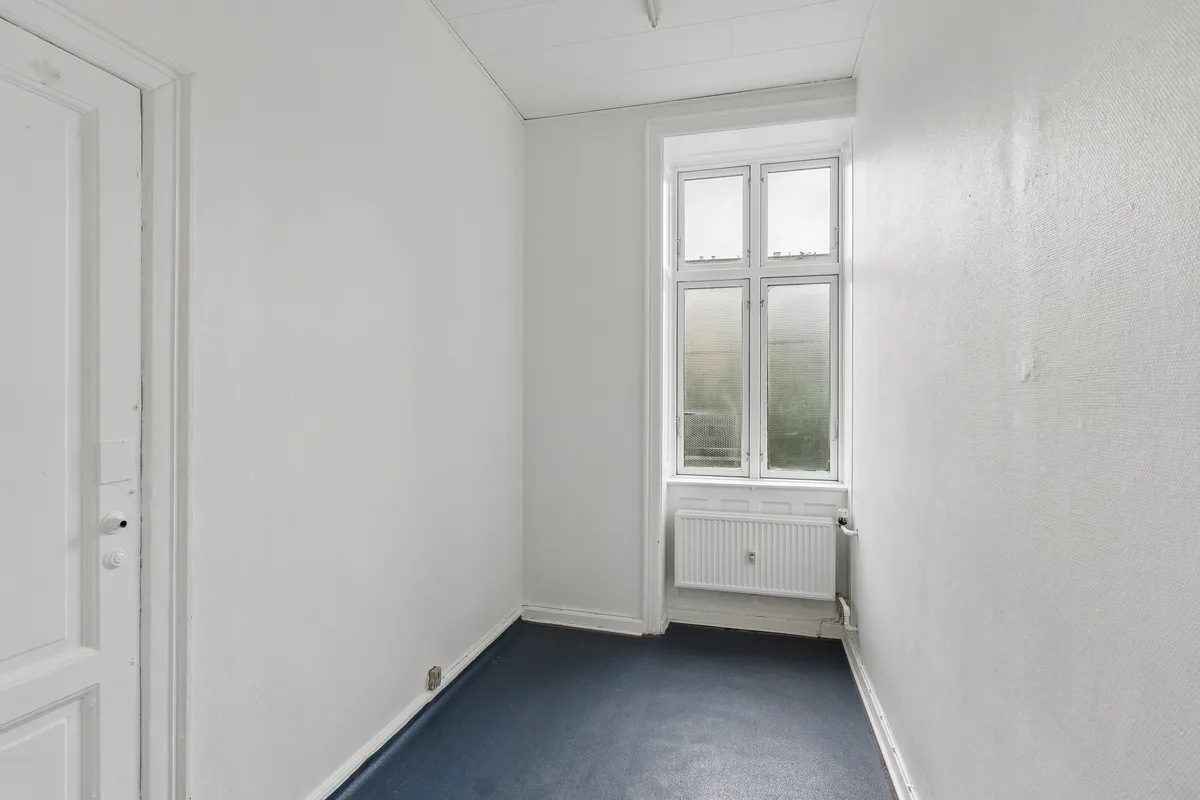 En tom rummelig værelse med hvide vægge og gulvtæppe, et vindue med dobbeltruder og radiator samt en hvid dør med to håndtag og et lås