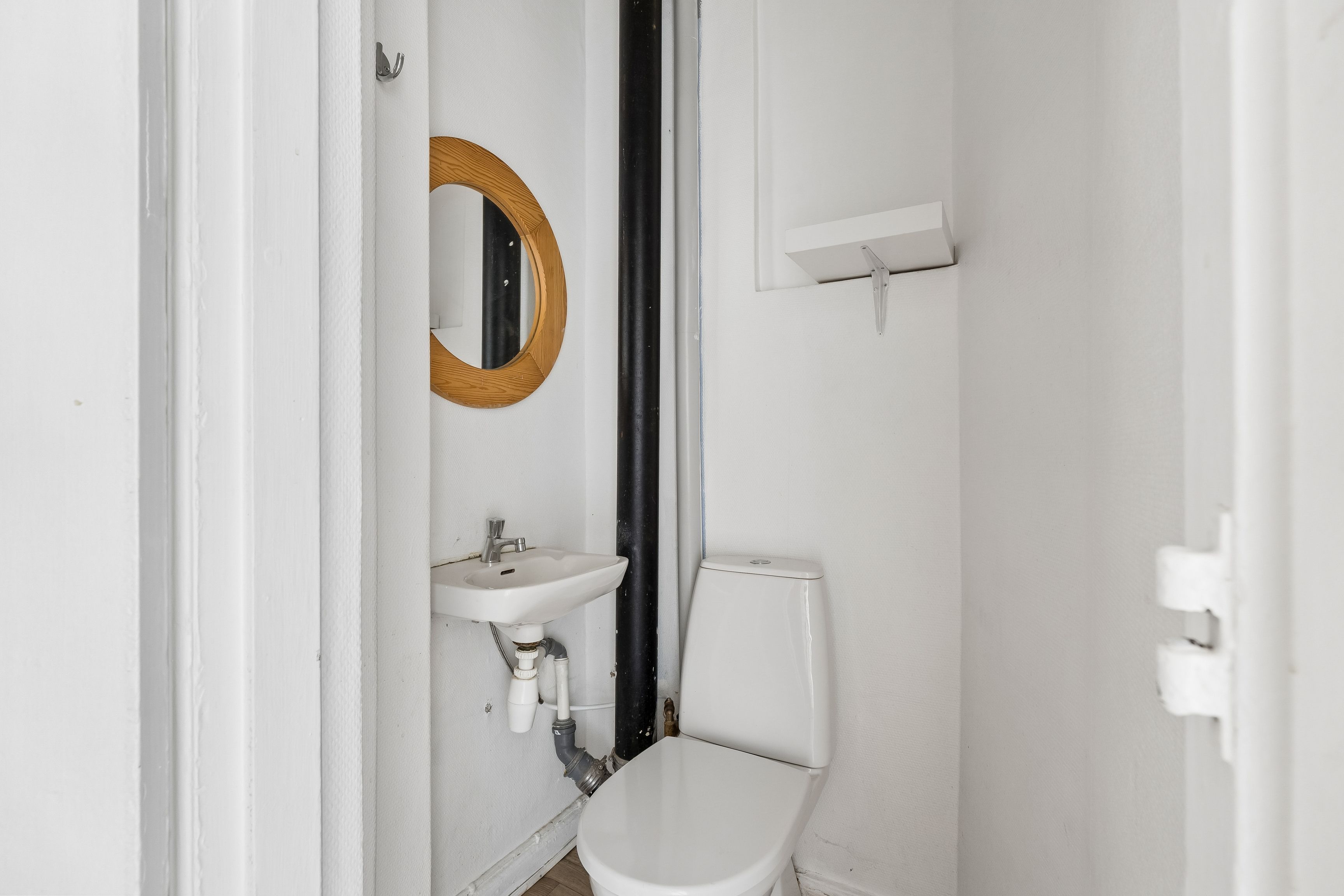 Hvidt badeværelse med toilet, håndvask og spejl på væggen og hængehylde med toiletpapir