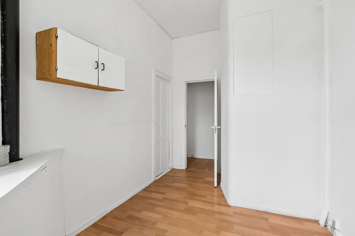 Lyst erhvervslejemål med store facadevinduer, hvidt vægmaleri, lyse trægulve og åben dør til et rum med spejl og lys.