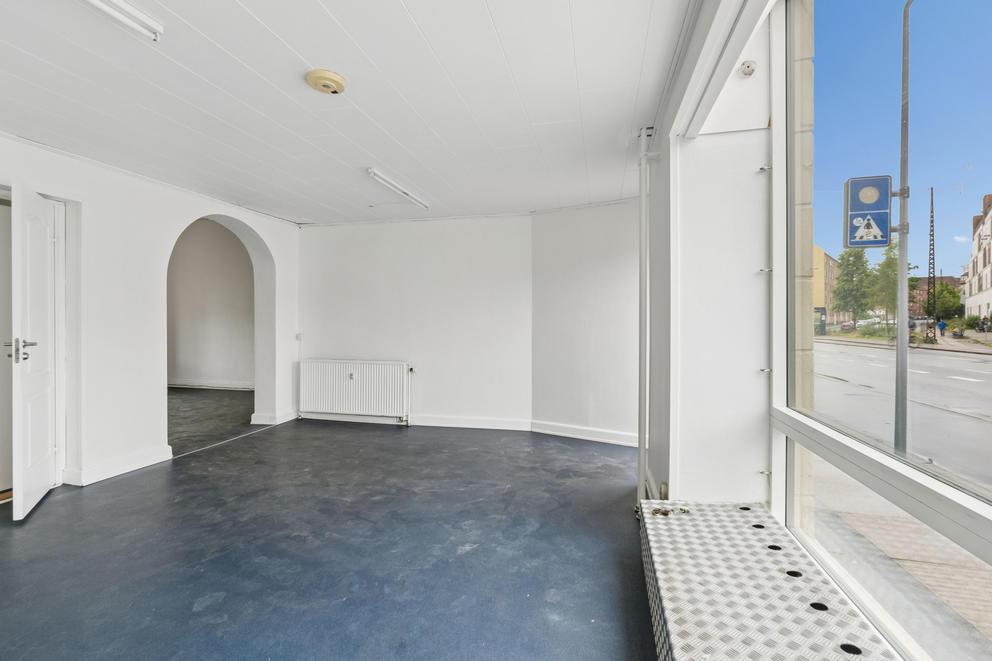 Store facadevinduer med glas og metal og en gulvkasse til udstilleskilt, hvide vægge og loft samt åben dør til et lysere rum med hvid ovn
