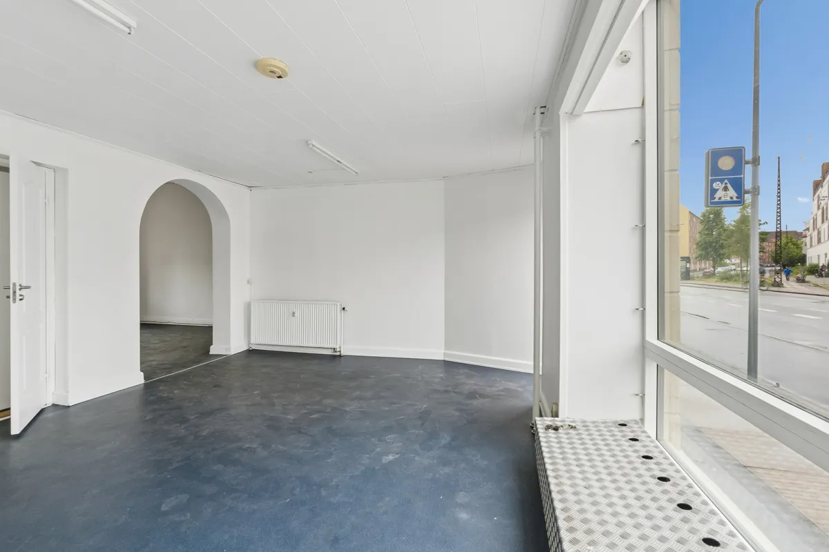 Store facadevinduer med glas og metal og en gulvkasse til udstilleskilt, hvide vægge og loft samt åben dør til et lysere rum med hvid ovn