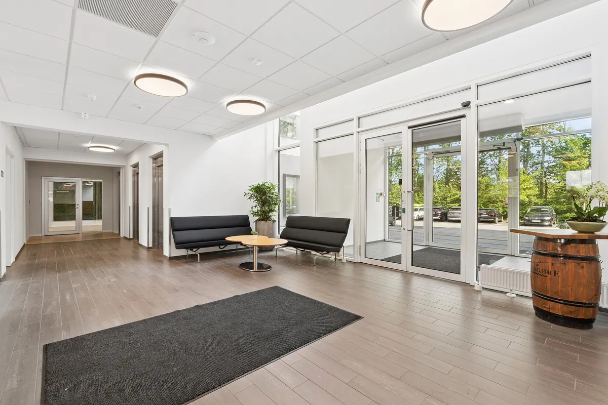 Moderne lobby med højt loft og glasdøre til gårdsplads. To sorte sofaer med højt gulv. Vinfat og plante på høj, trægulv. Mødecenter og kontorer til leje i Vallensbæk Strand