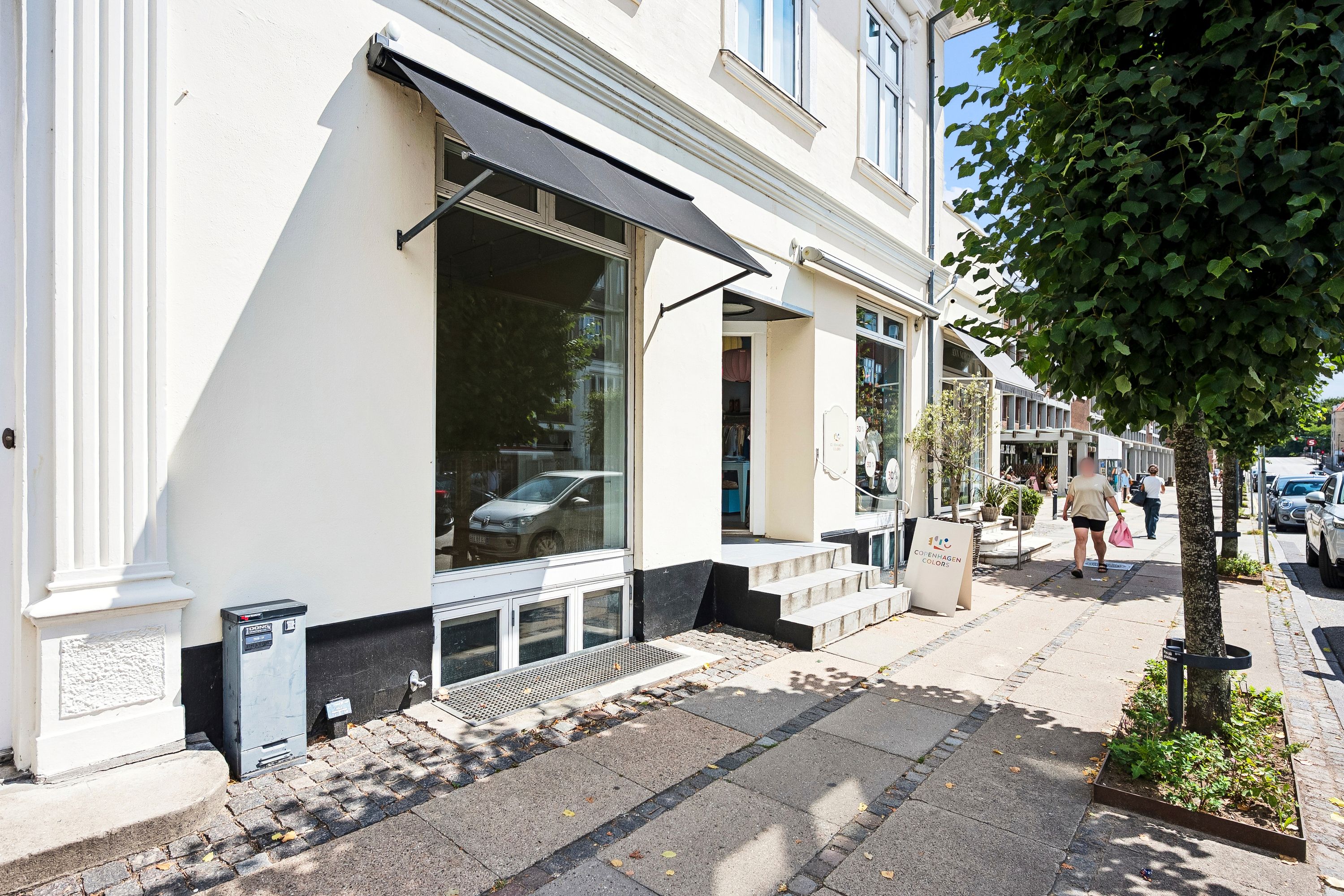 Moderne butik med glasvinduer og skrånende fortovskant. Solbeskyttelse og træplantning sammen med biler langs vejen. Mennesker med indkøbsposer går på fortovet.