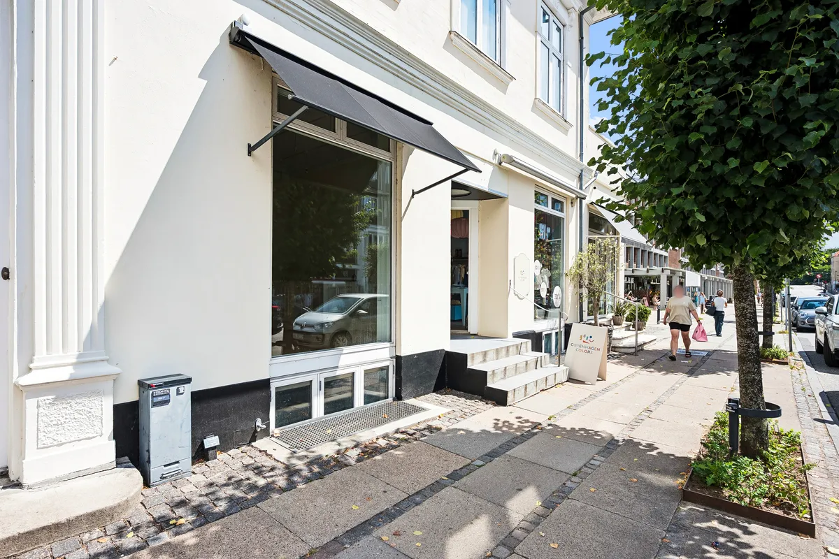 Moderne butik med glasvinduer og skrånende fortovskant. Solbeskyttelse og træplantning sammen med biler langs vejen. Mennesker med indkøbsposer går på fortovet.