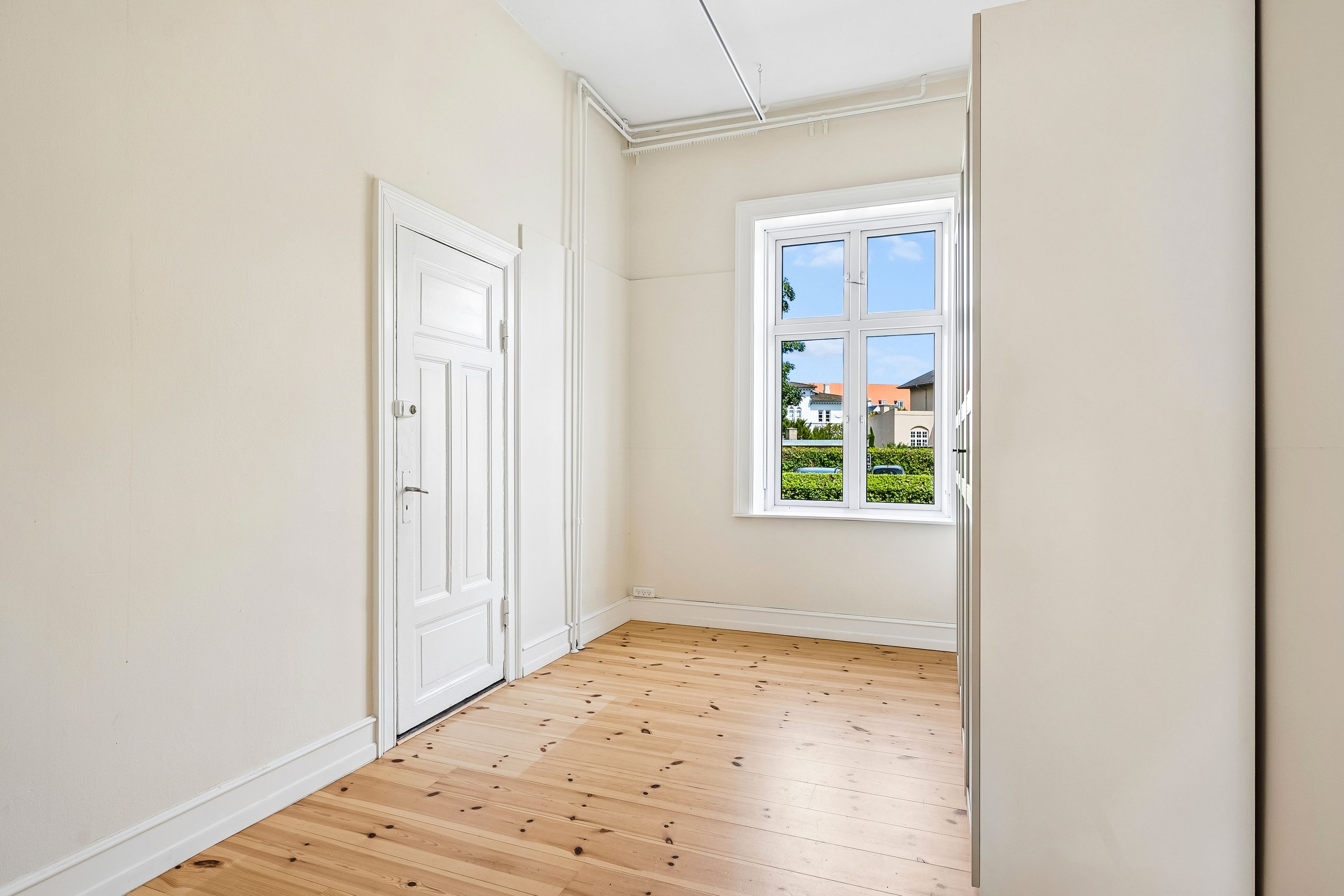 Et rum med hvidt vægmaleri og lysegrå loft. Et hvidt dørepar og et vindue med udsigt til grønne planter og bygninger. Gulvet er lavet af træ