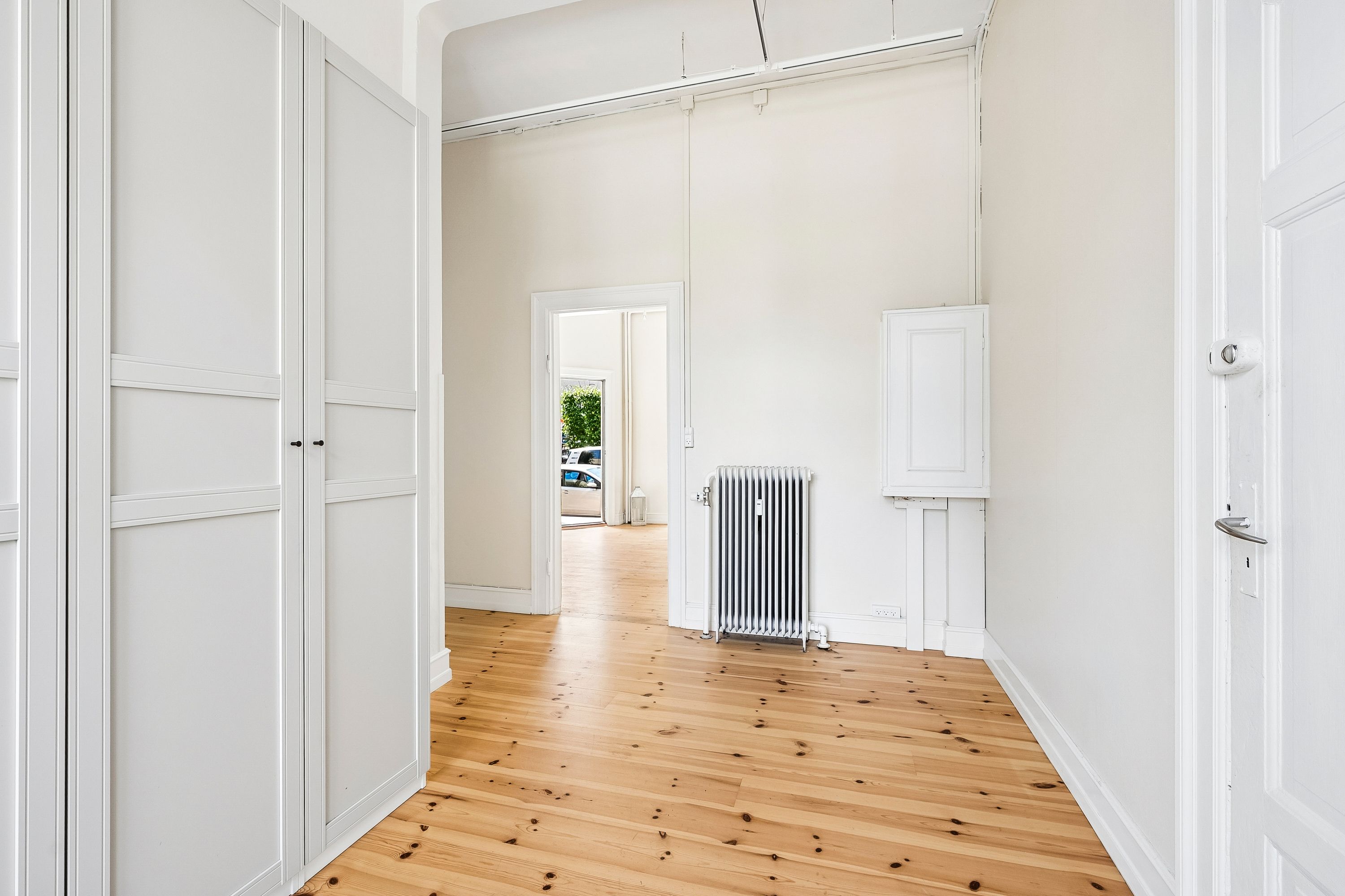 To hvide kabinetter med dobbeltdøre og hvid radiator samt to hvide døre i en tom rum. Gulvet er lavet af træ med mørke prikker